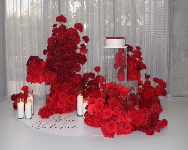 Red flowers arranged on a table with candles and a decorative white circular sign with red writing that says "Kevin Valeria" in front of a sheer curtain.