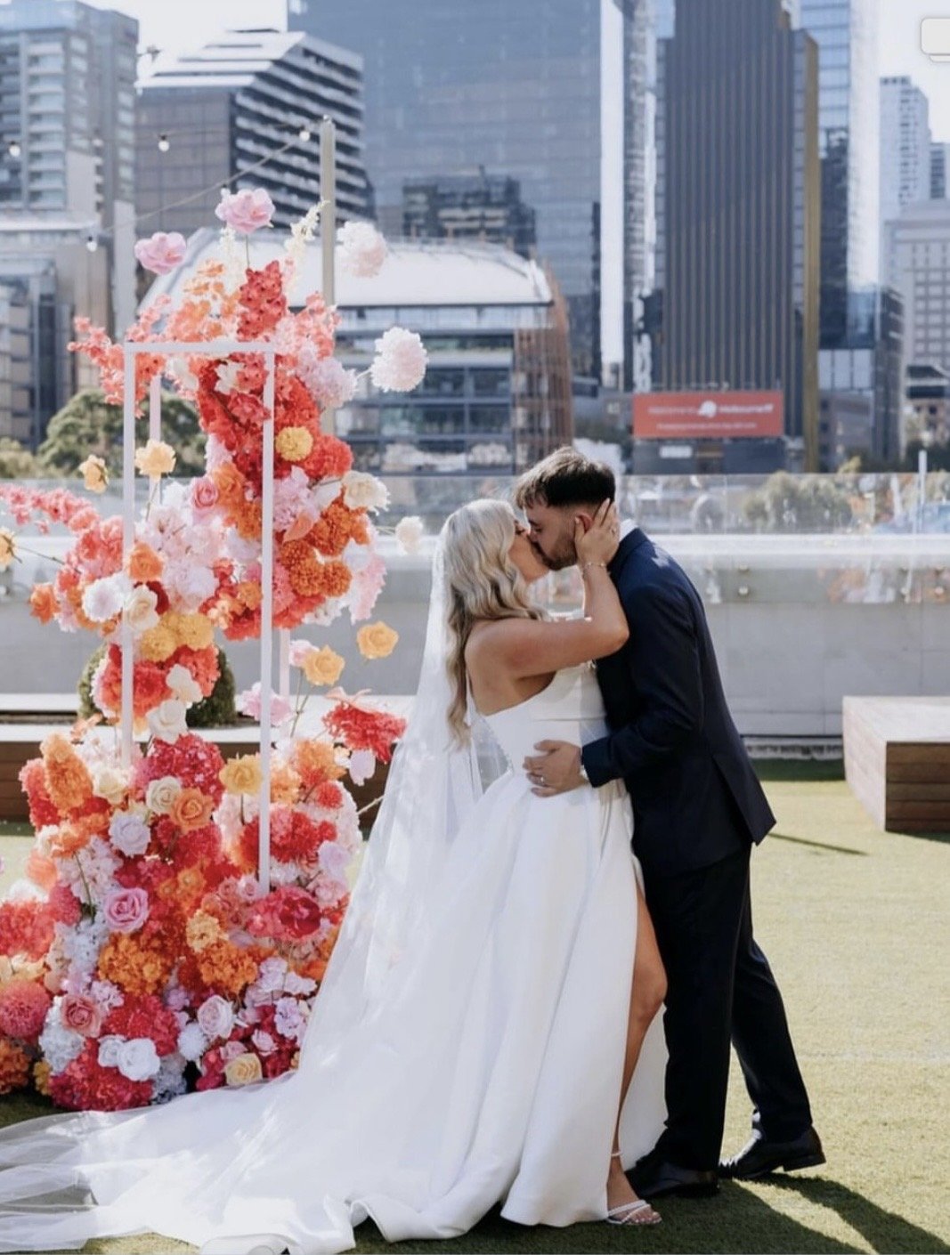 A bride and groom kiss at their wedding ceremony in an outdoor urban setting with tall buildings in the background. The bride is wearing a white gown with a train and the groom is in a dark suit. There is a tall floral arch decorated with pink, orang