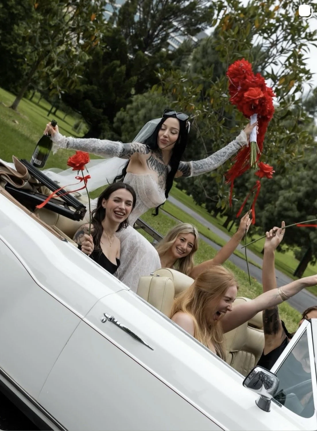 Women celebrating in a white convertible car, one woman in a wedding dress holding a bouquet and a bottle of champagne, others smiling and waving, outdoors in a park with trees.