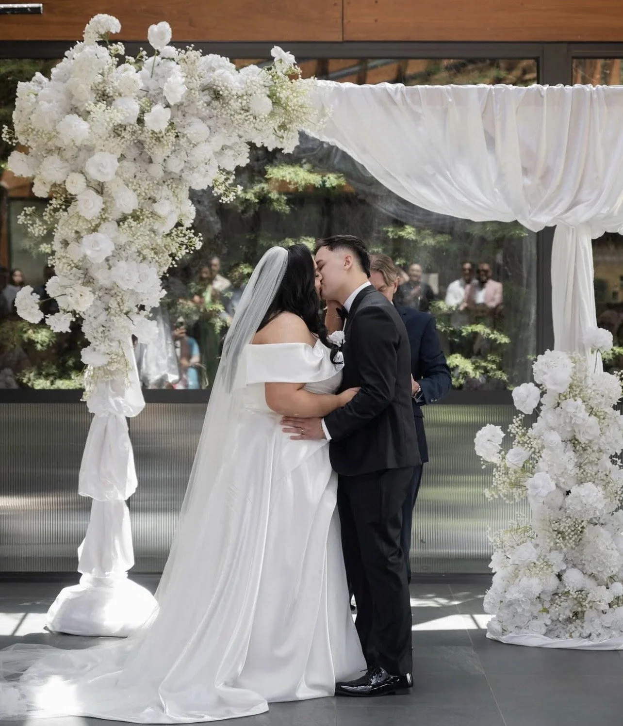 A bride and groom kiss during their wedding ceremony under a white floral and fabric arch.