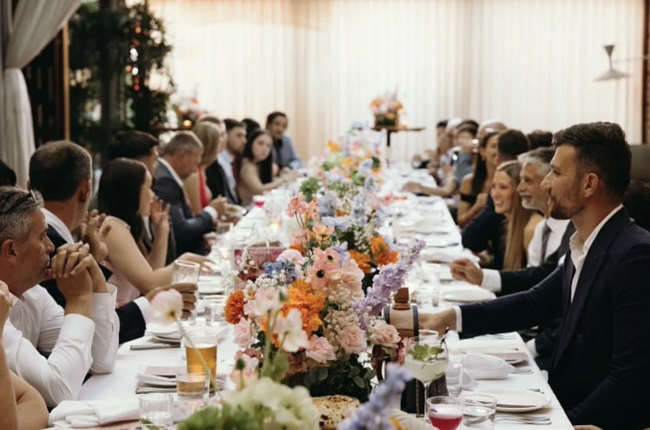 A long banquet table decorated with colorful flowers, with guests dressed in formal attire seated on both sides, celebrating in a well-lit indoor setting with cream-colored curtains.