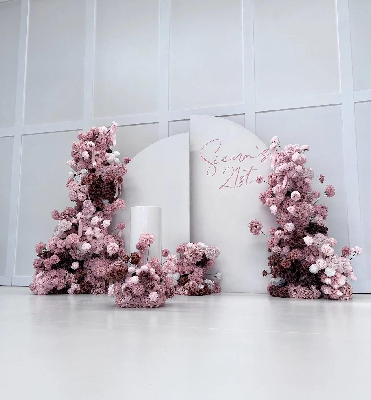 Floral arrangement of pink and white flowers surrounding a white backdrop with the text "Siena's 21st" in pink cursive. The background is a plain white wall and floor.