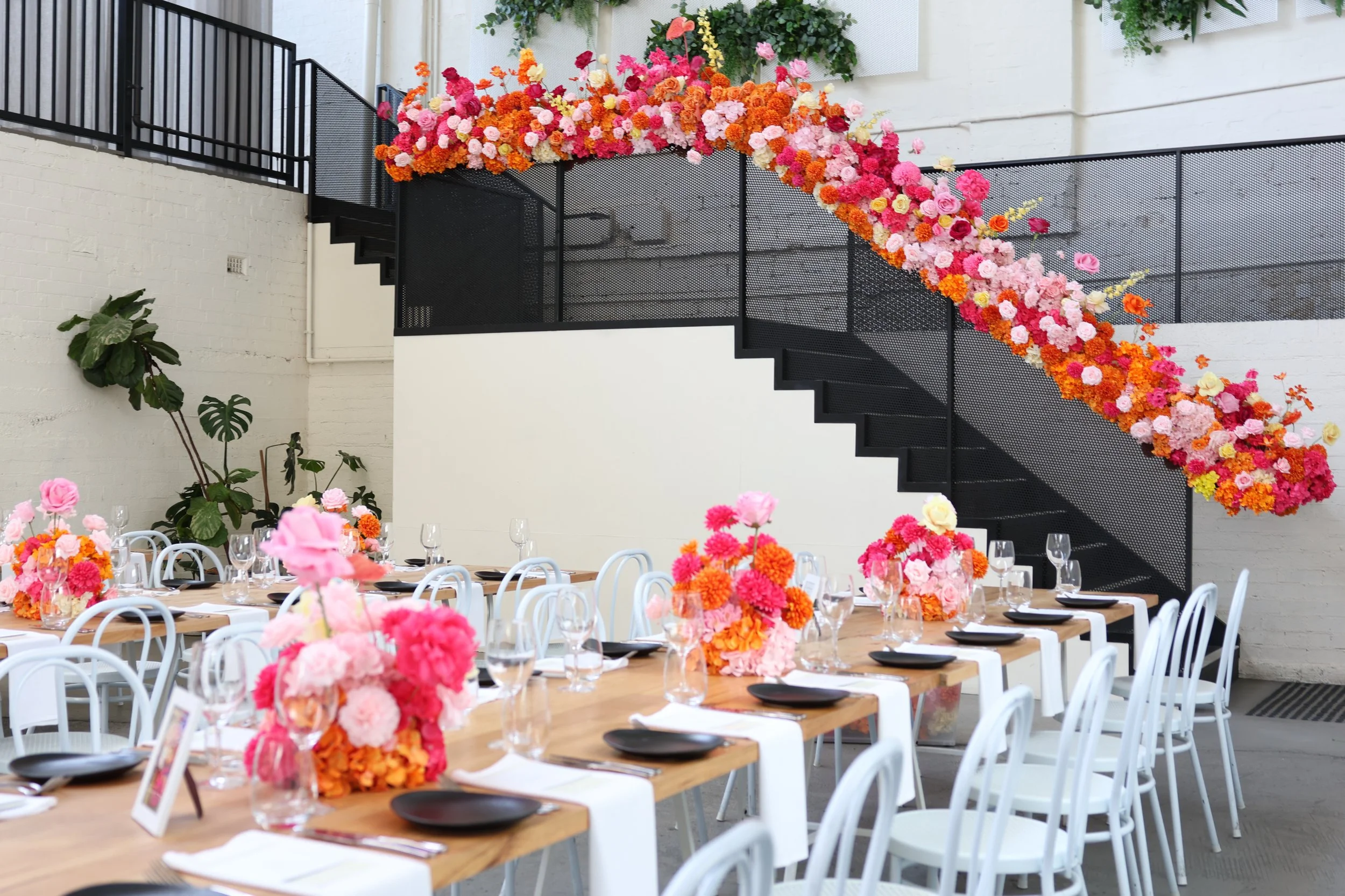 Indoor event space decorated with vibrant pink, orange, and peach flower arrangements on tables and a staircase with a floral display along the black railing.