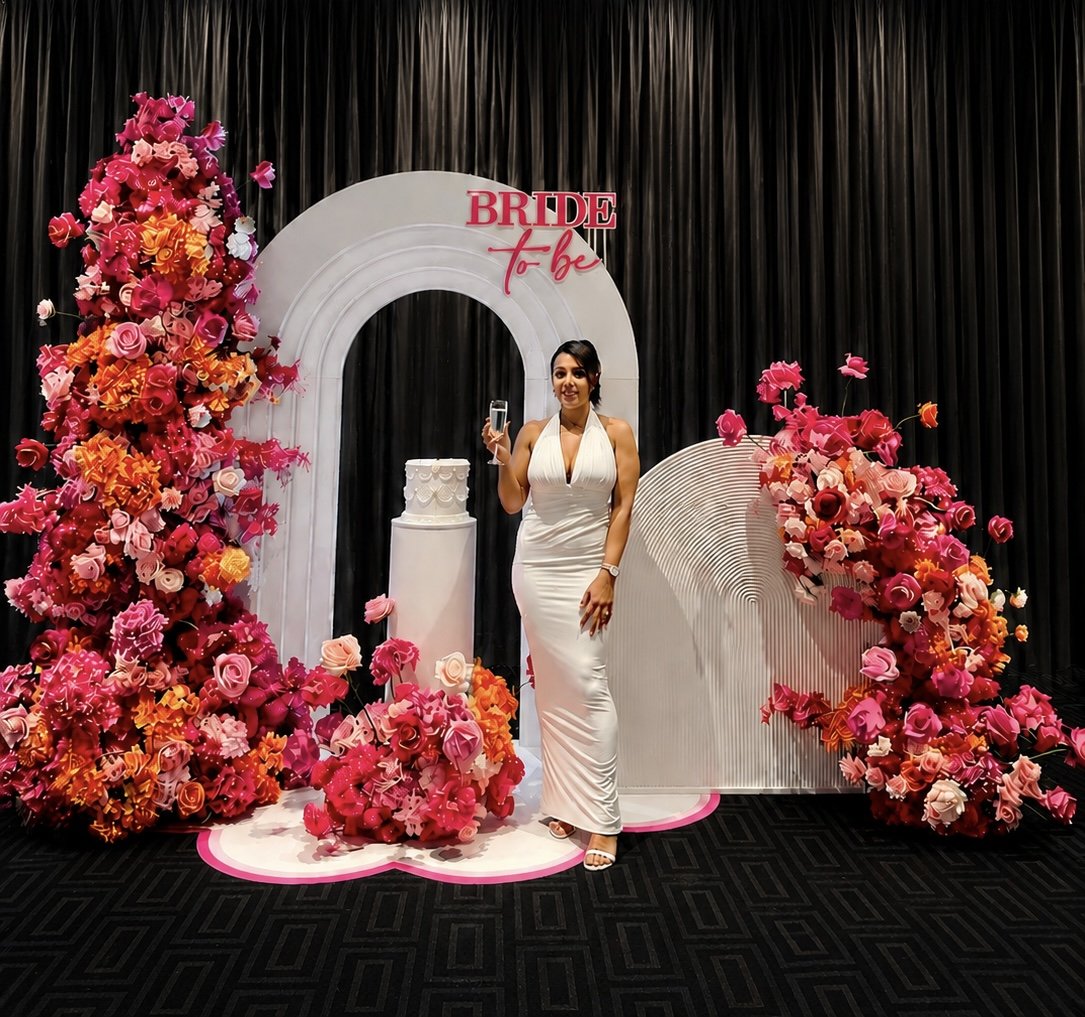 A woman in a white gown holding a glass of champagne, standing in front of a wedding-themed backdrop with pink and orange flowers, a white arch, and a sign that says 'Bride to be'.