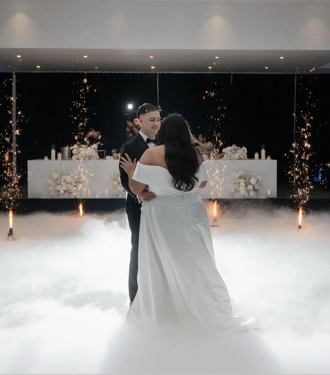 A bride and groom sharing their first dance at a wedding reception, with sparklers and fog effects in the background.