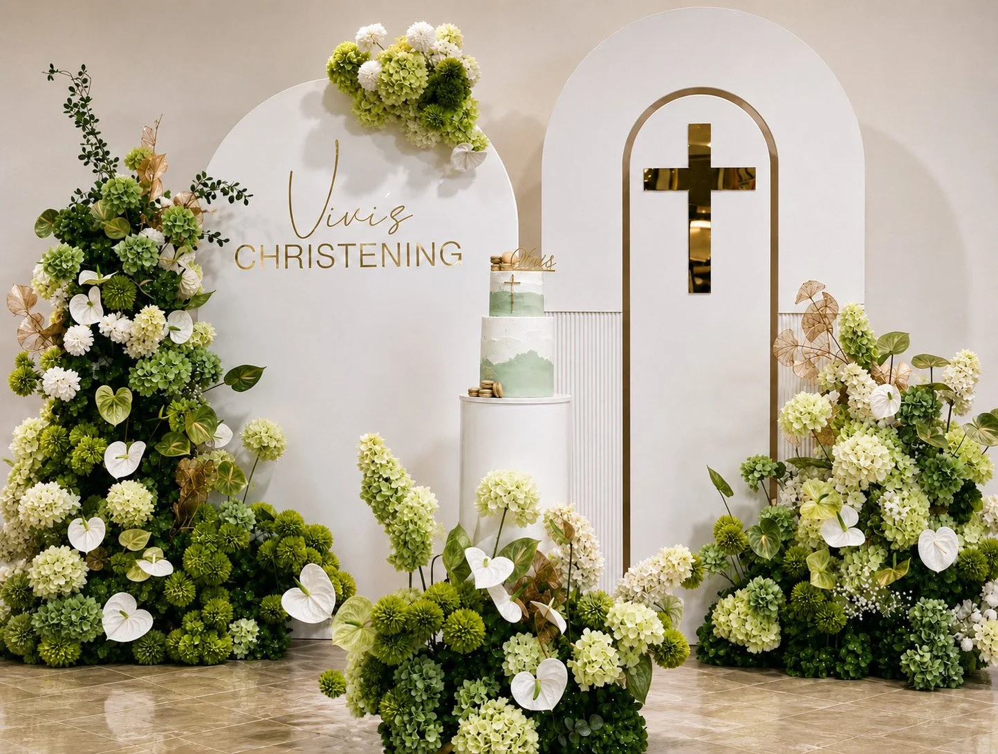 Elegant wedding altar decorated with white and green flowers, including hydrangeas and anthuriums, surrounding a white cake with green watercolor design and gold accents, and a minimalist backdrop with a gold cross and the names 'Vinis Christening' i