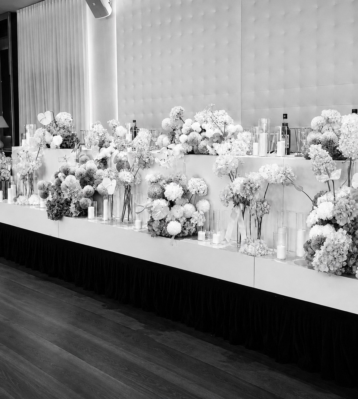 A black and white image of a decorated table with numerous floral arrangements and candles, set against a padded wall at an event or celebration.