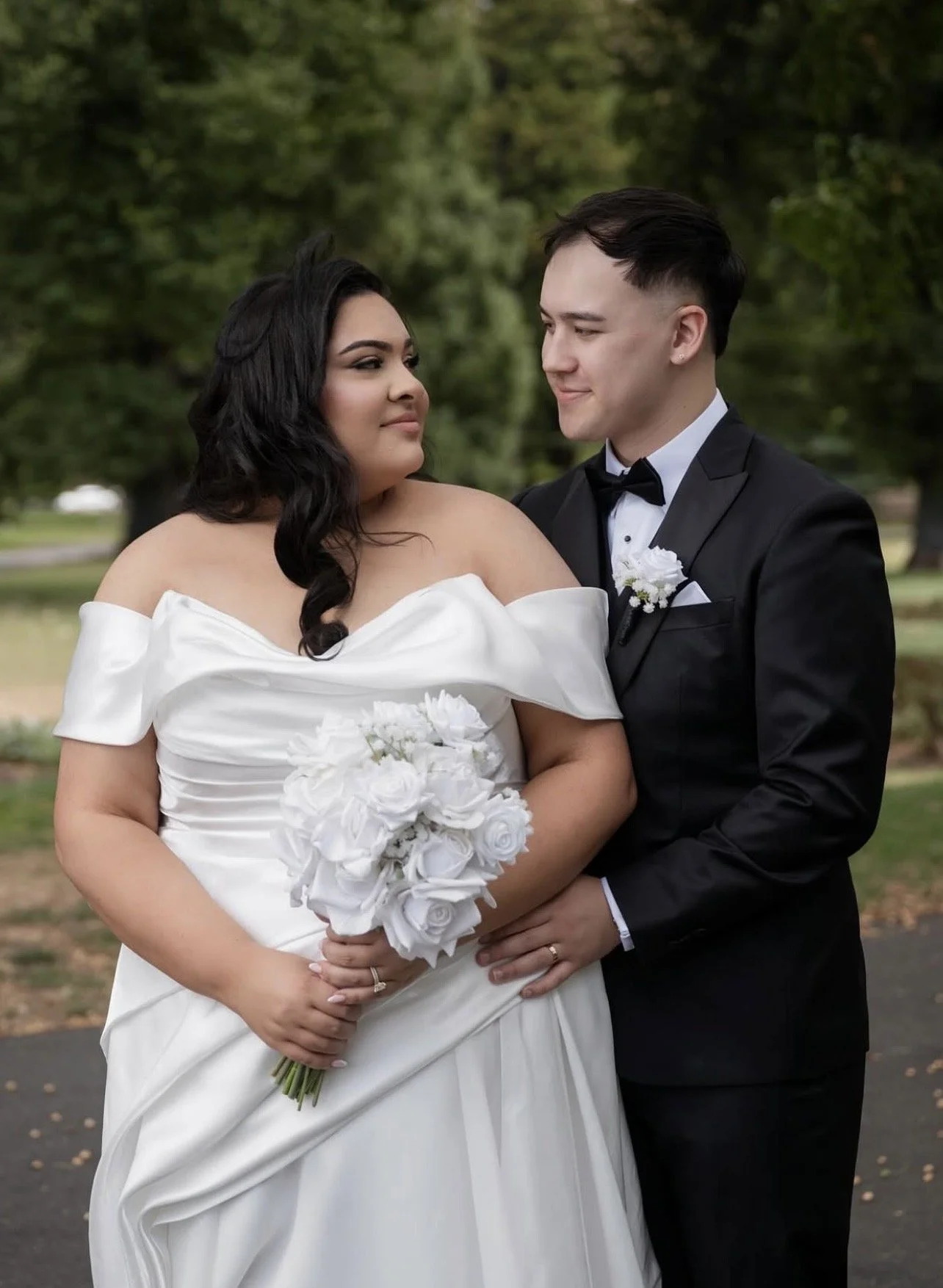 A newlywed couple standing outdoors, the bride in an off-shoulder white wedding gown holding a bouquet of white roses, and the groom in a black tuxedo with a bow tie, with trees in the background.