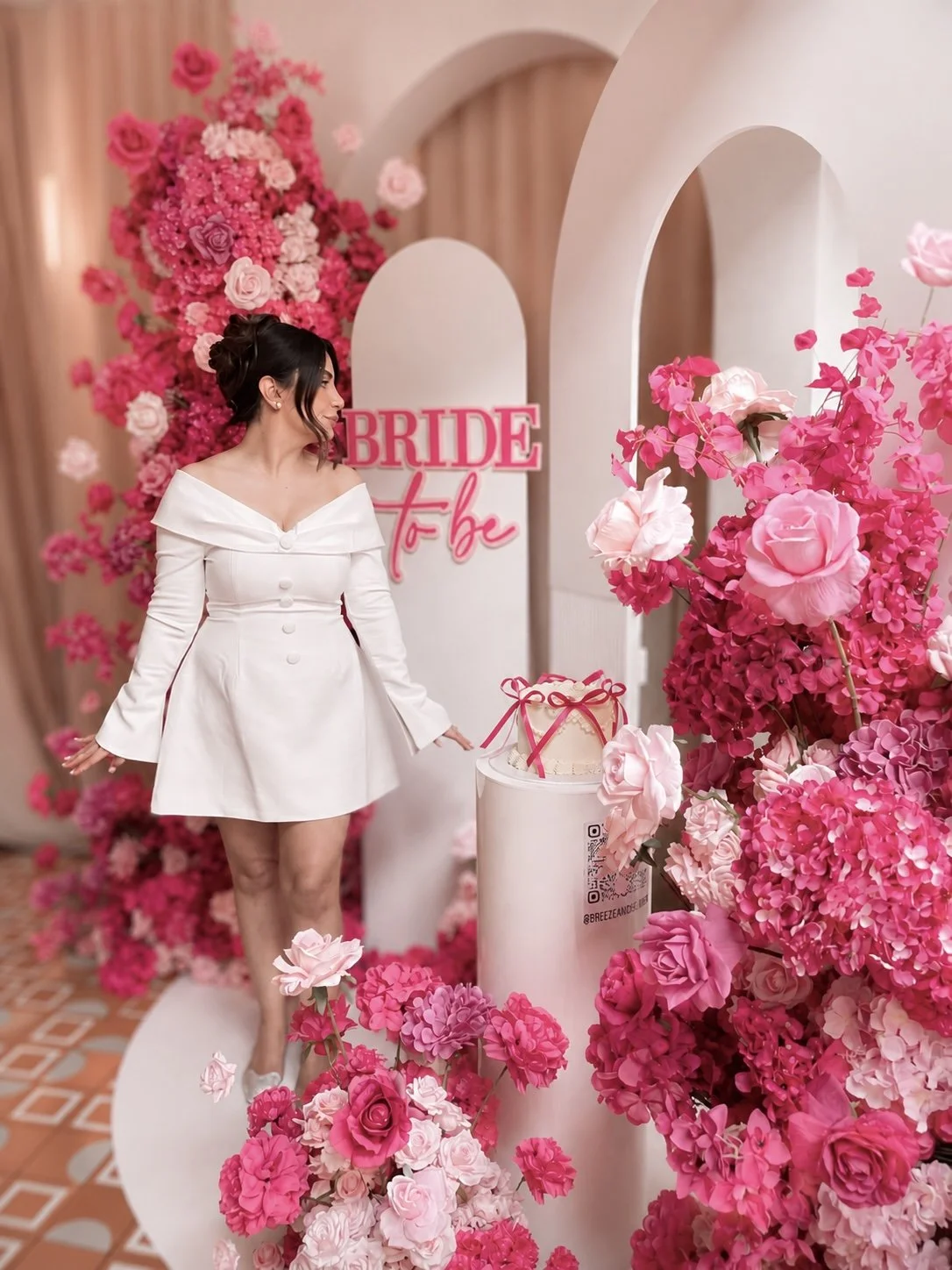 A woman in a white dress standing among pink and red flowers, with a sign behind her that says 'Bride To Be' in pink letters.