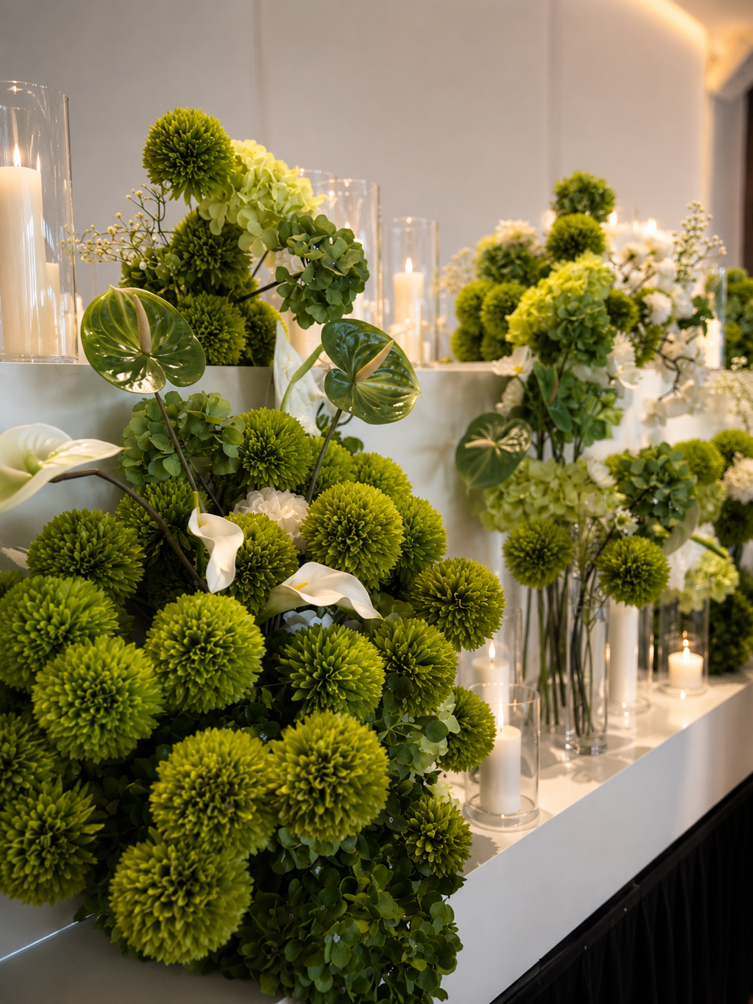 Arranged green and white floral centerpiece with candles in tall glass holders on a white table.