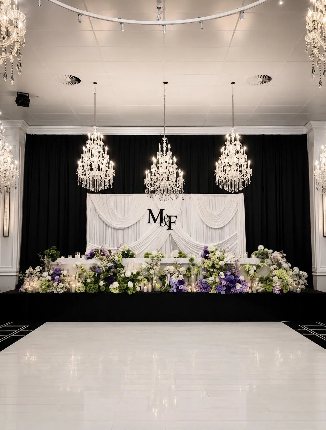 Elegant wedding or event stage with white drapery, floral arrangements in purple, white, and green, lit candles, and crystal chandeliers, with initials "M & F" displayed in the center.