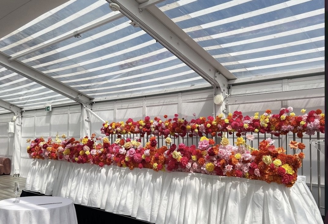 A long table decorated with a white tablecloth and filled with colorful flowers inside a glass-roofed venue.
