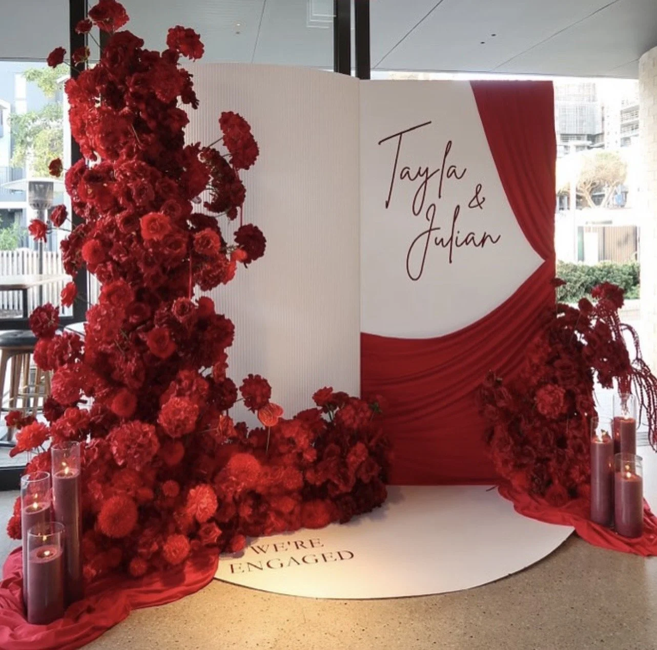 Red floral wedding display with a backdrop featuring the names 'Tayla & Julian,' surrounded by red flowers and candles, with the words 'We're Engaged' on the floor.
