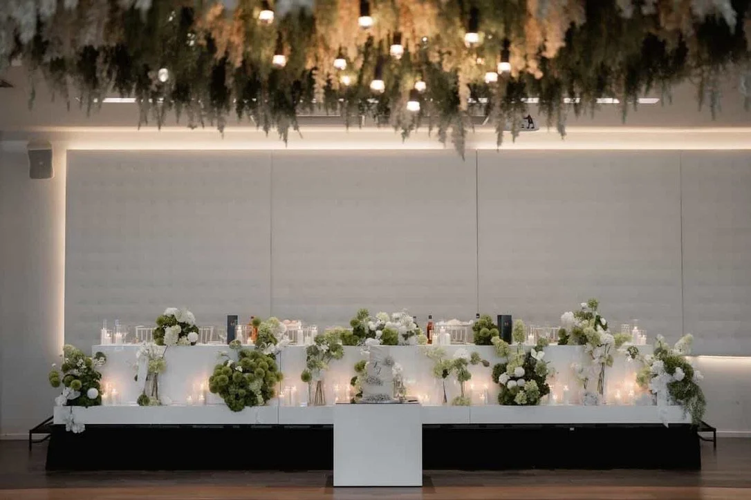 Elegant event setup with a long white table adorned with white and green floral arrangements, candles, and bottles, against a plain wall with minimal lighting.