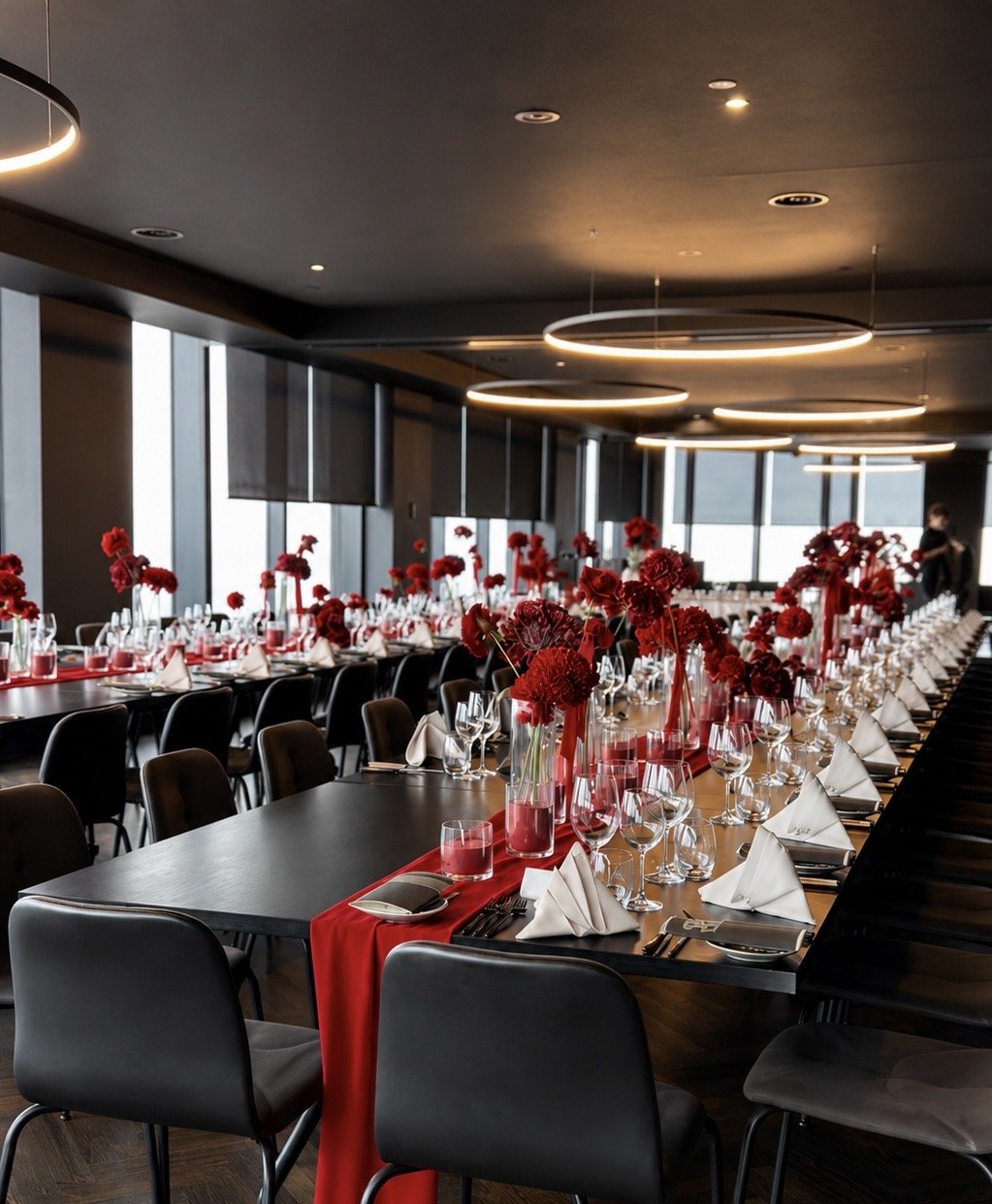 Elegant dining room decorated with red flowers, tableware, and white napkins for an event, with large windows and modern circular ceiling lights.