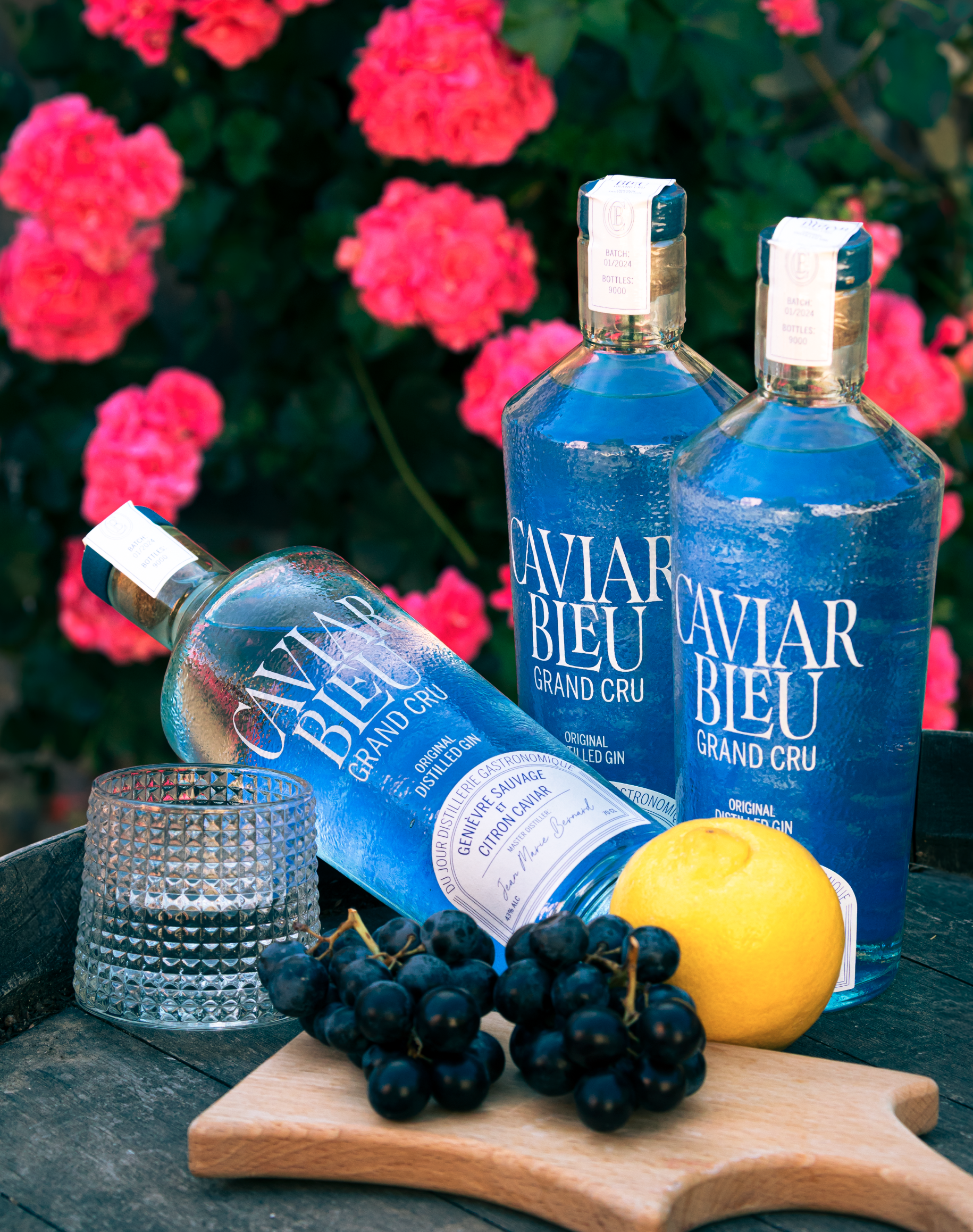 Deux bouteilles de gin bleu clair, une grappe de raisins noirs, un citron jaune, une coupe à whisky en verre gravé, des fleurs roses en arrière-plan, et un plateau en bois.