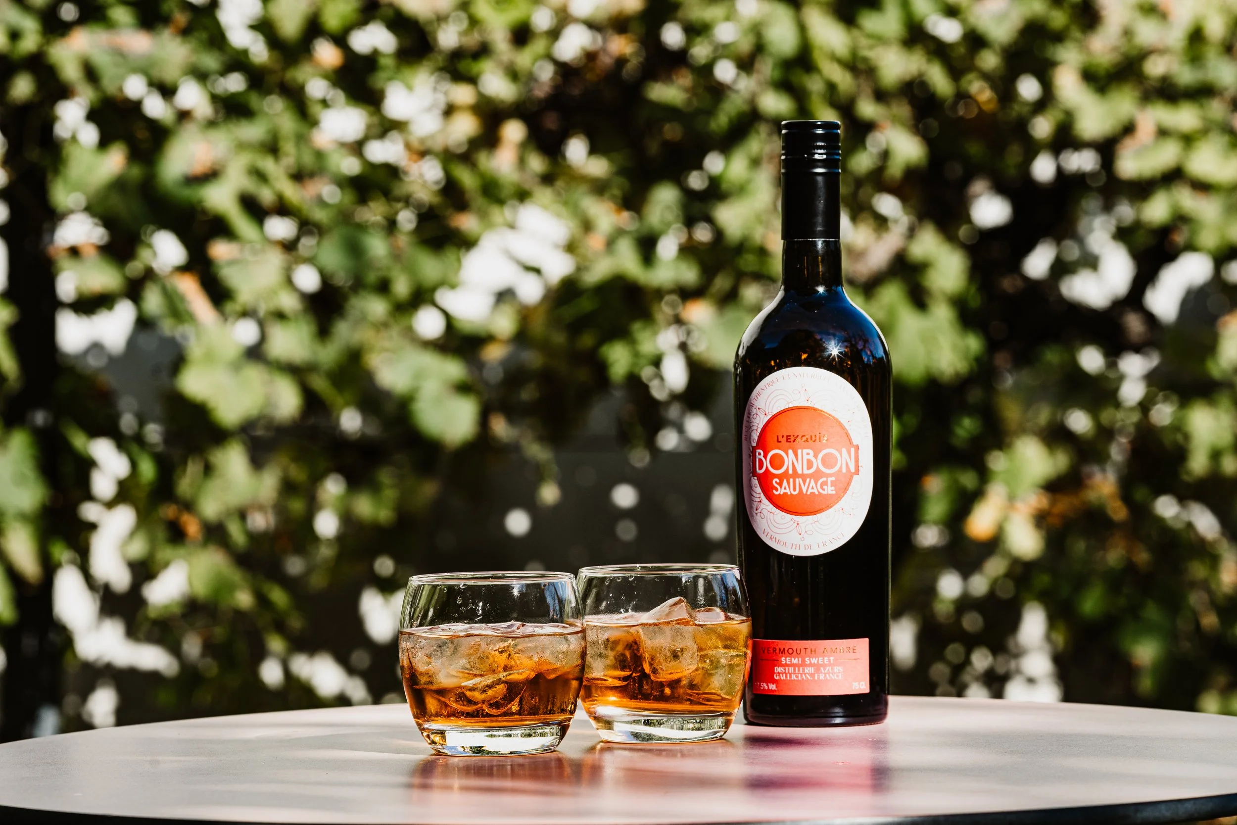 Une bouteille de vermouth Ambré Bonbon Sauvage et deux verres avec une boisson glacée, sur une table en extérieur avec un fond de feuillage vert.