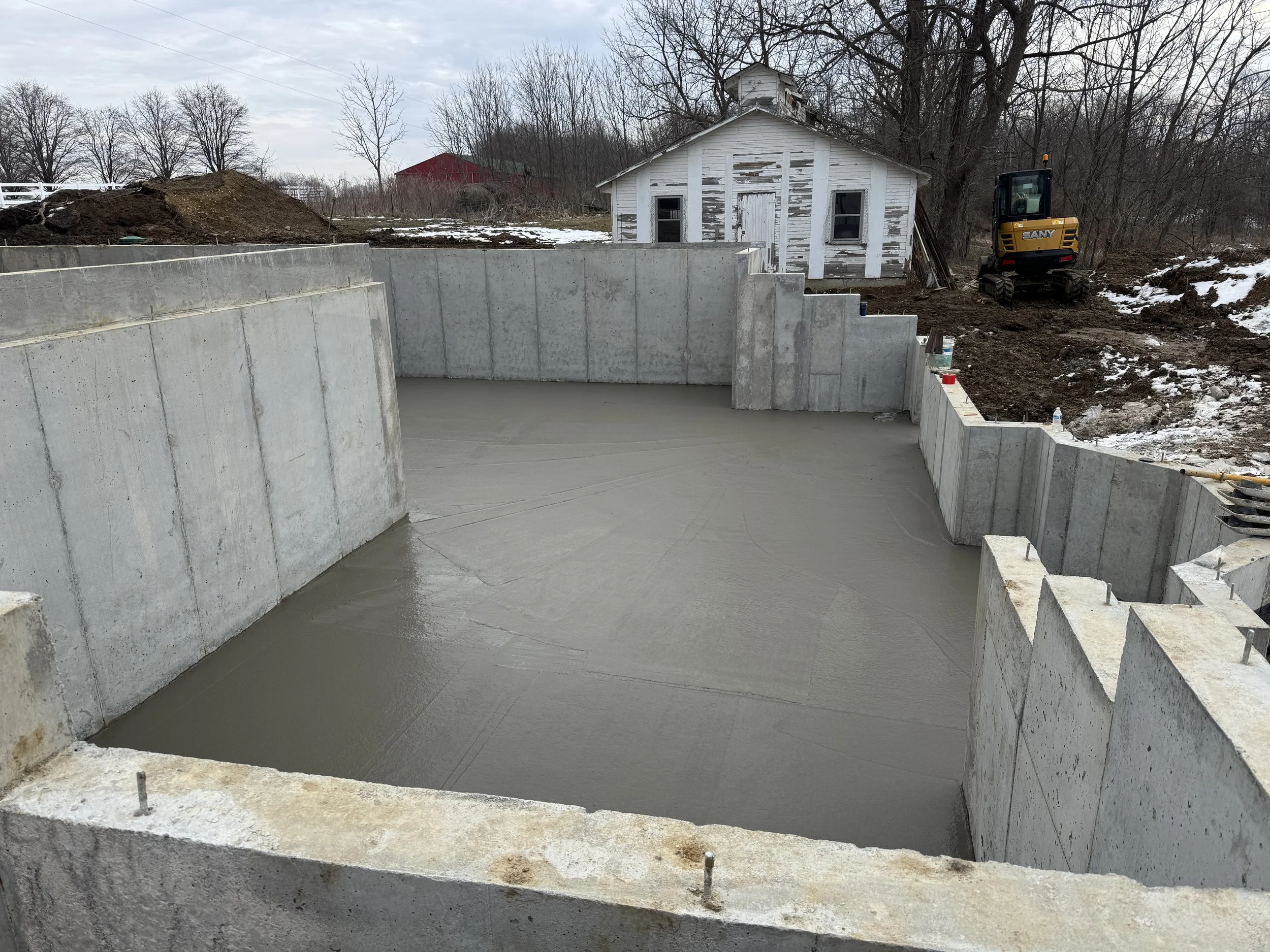 Construction site showing a partially built concrete foundation with a concrete slab that was just poured and construction equipment in the background, along with trees and snow as this was in December.