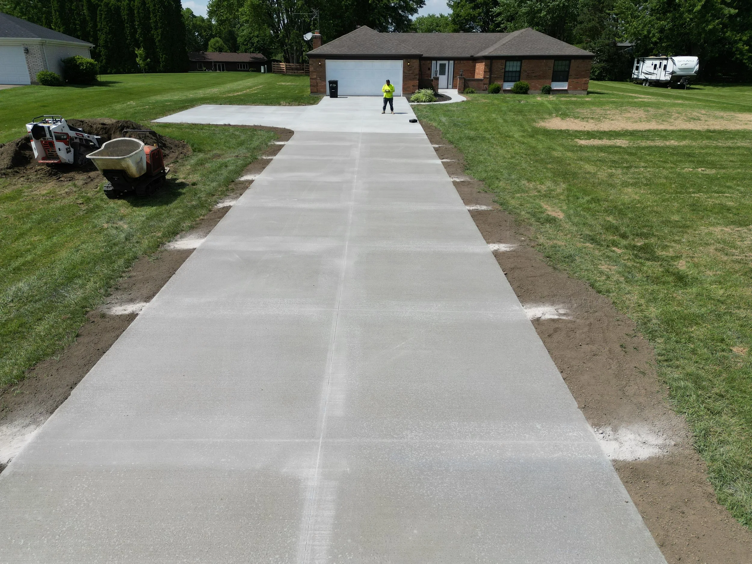 Newly poured concrete driveway leading to a brick house, with construction tools and equipment nearby as saw cuts are being cut in and the jobsite is being cleaned up before completion.