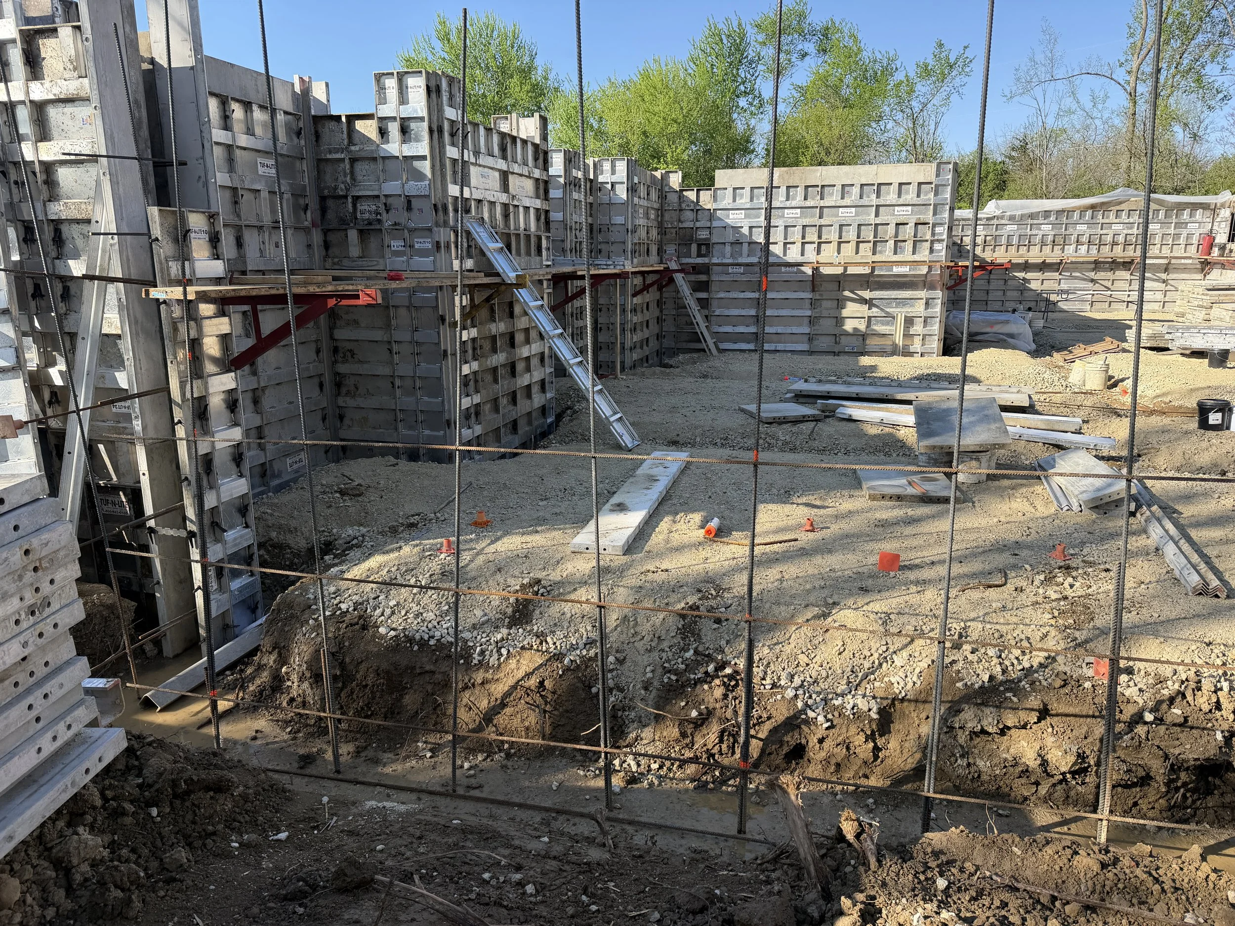 Construction site with concrete form walls and steel rebar, ladders, and construction materials, with trees in the background and a clear blue sky.