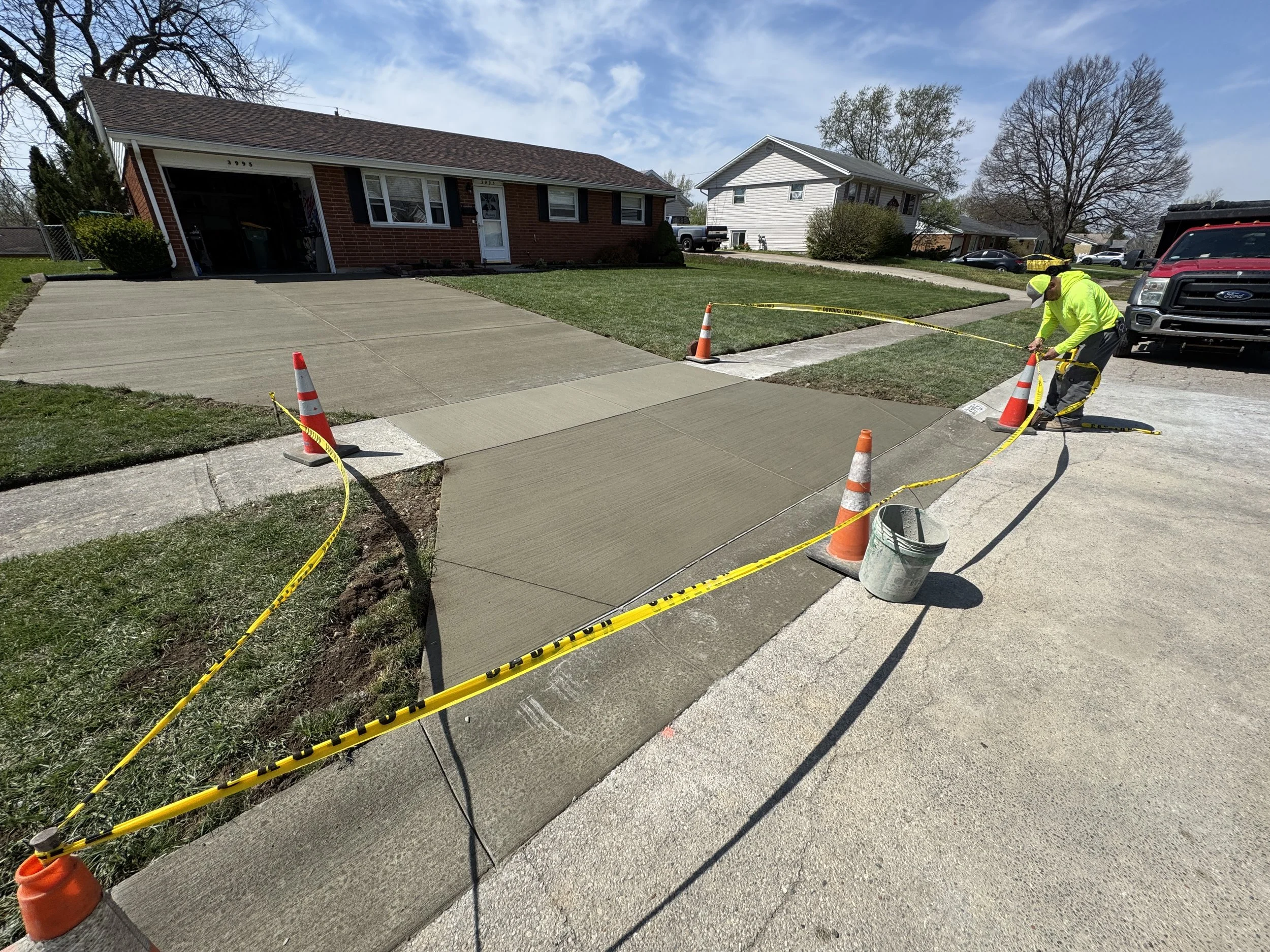 A worker is finishing freshly poured concrete on a sidewalk in a residential neighborhood, with orange cones and caution tape marking the area.