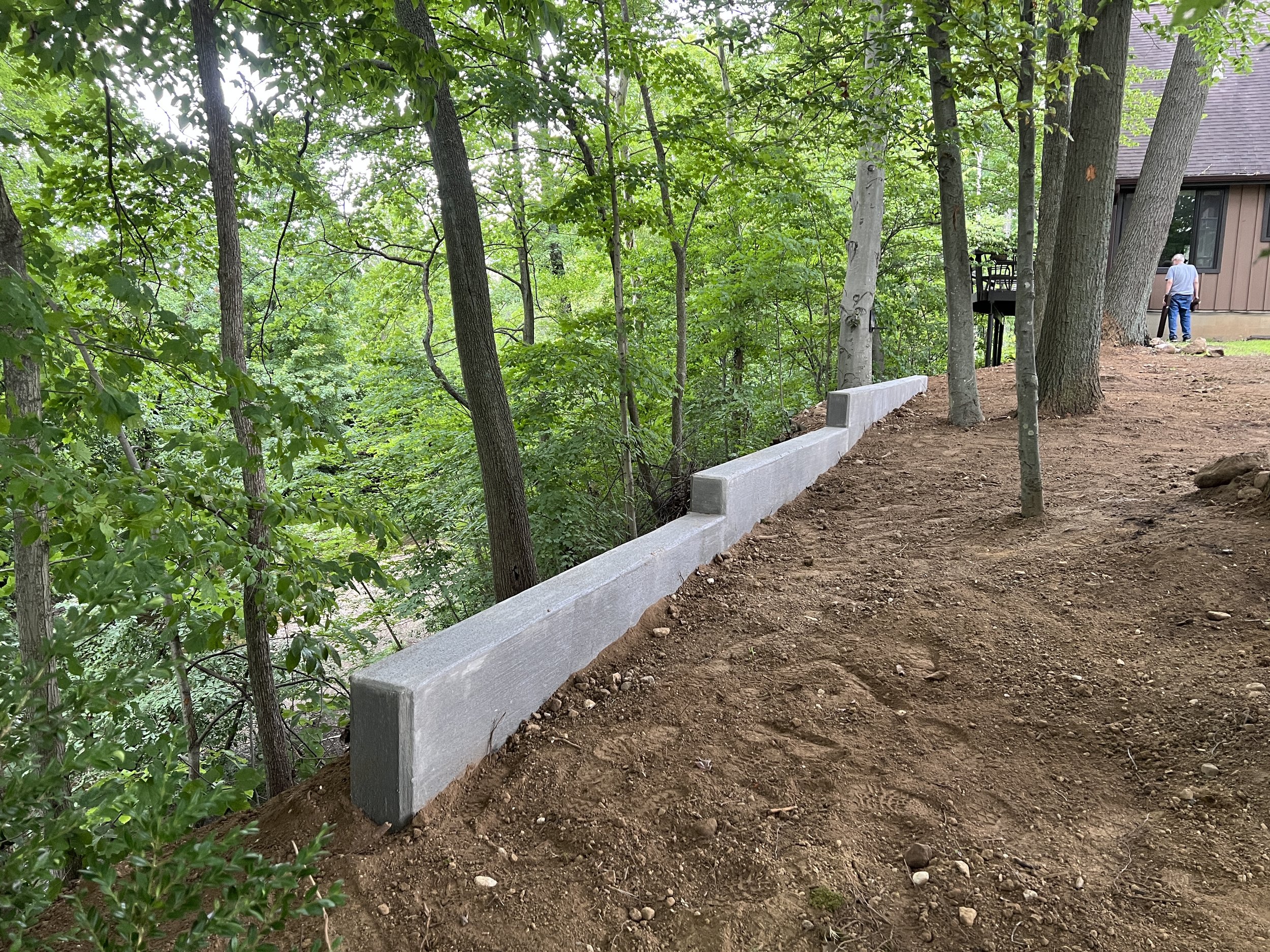 A backyard with trees and a house in the background. There is a concrete retaining wall along the dirt ground, which was just finished landscaped after construction.