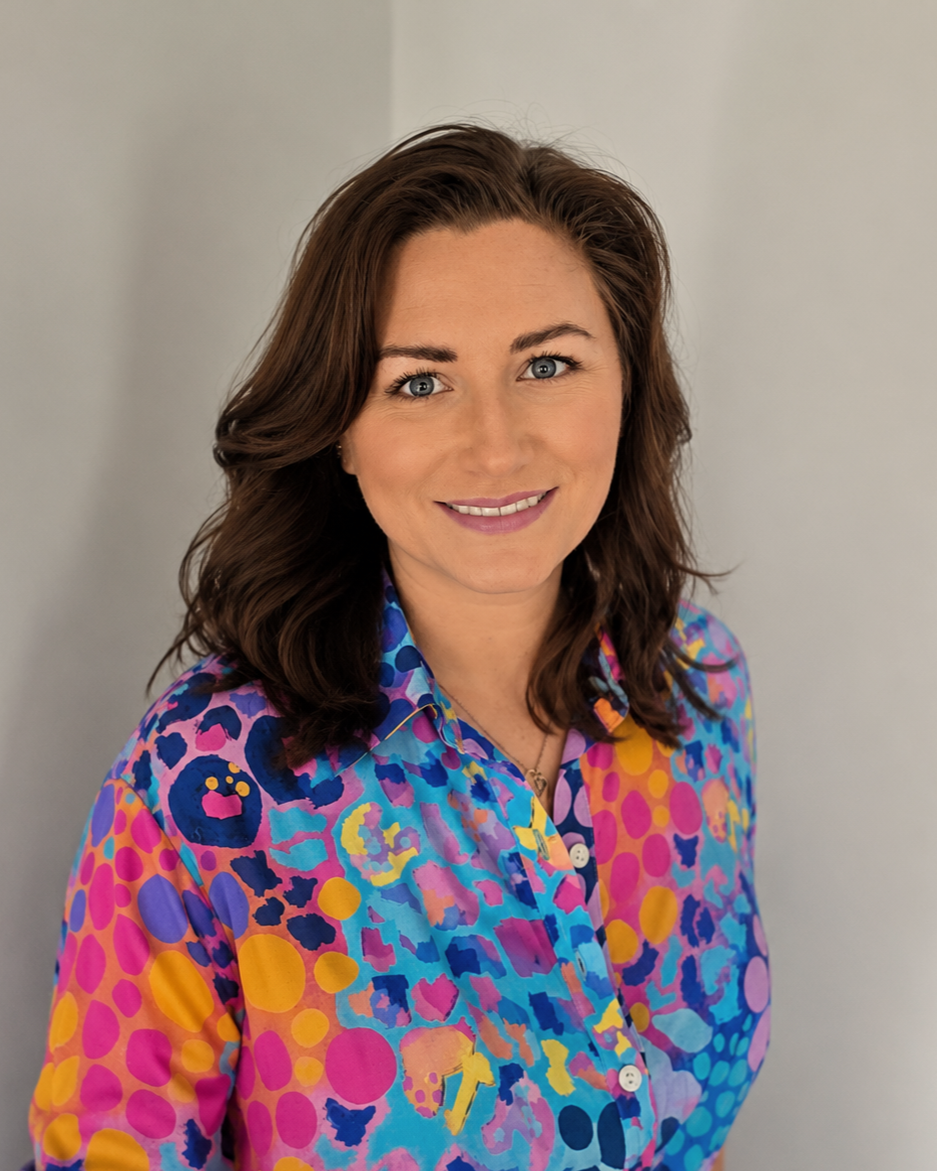 A woman with shoulder-length brown hair and blue eyes, wearing a colorful, patterned shirt with pink, blue, purple, and orange abstract designs, smiling at the camera against a plain gray background.
