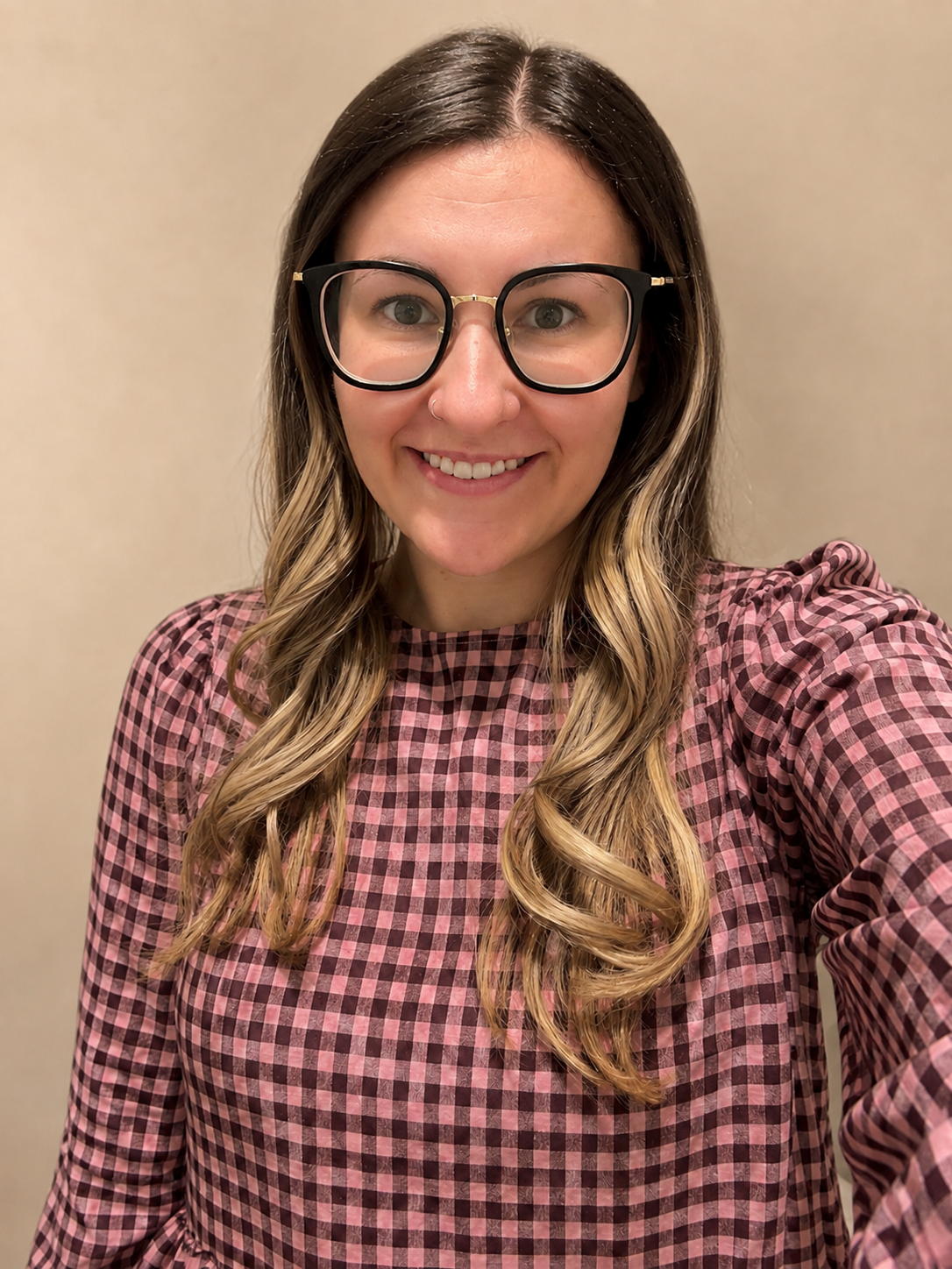 A woman with long wavy hair, wearing black glasses, and a pink checkered top, smiling at the camera indoors.
