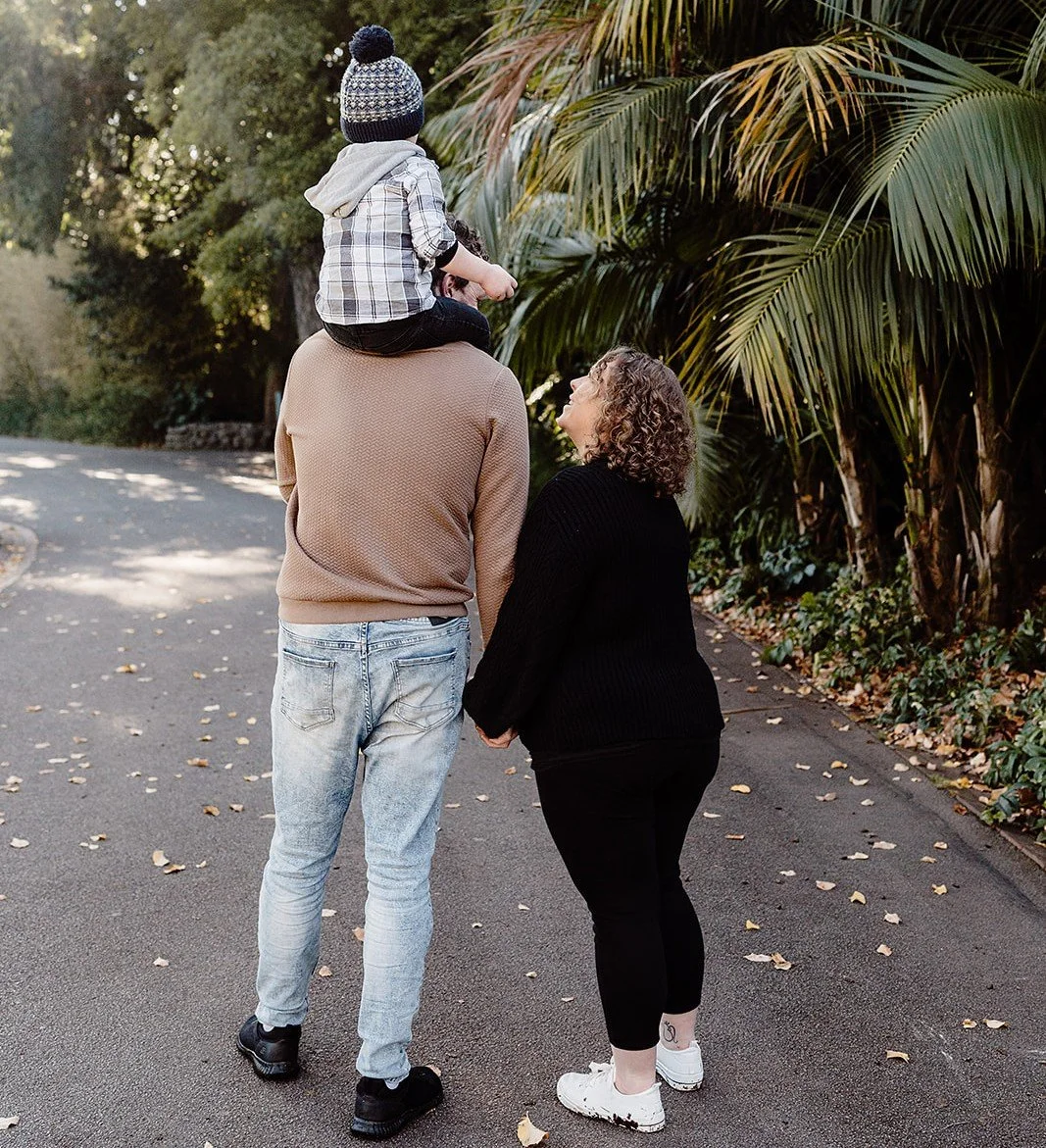 A family of three, a man, woman, and a child, walking and holding hands on a paved path surrounded by green tropical plants and trees, with the man carrying the child on his shoulders.