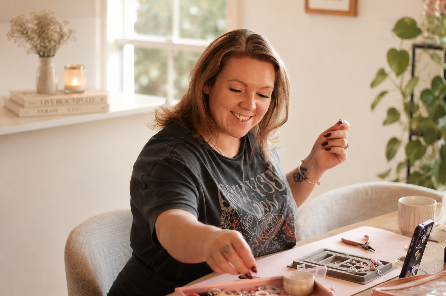 A woman with shoulder-length reddish hair smiling while sitting at a table, engaging in jewelry making with various jewelry supplies and tools around her.
