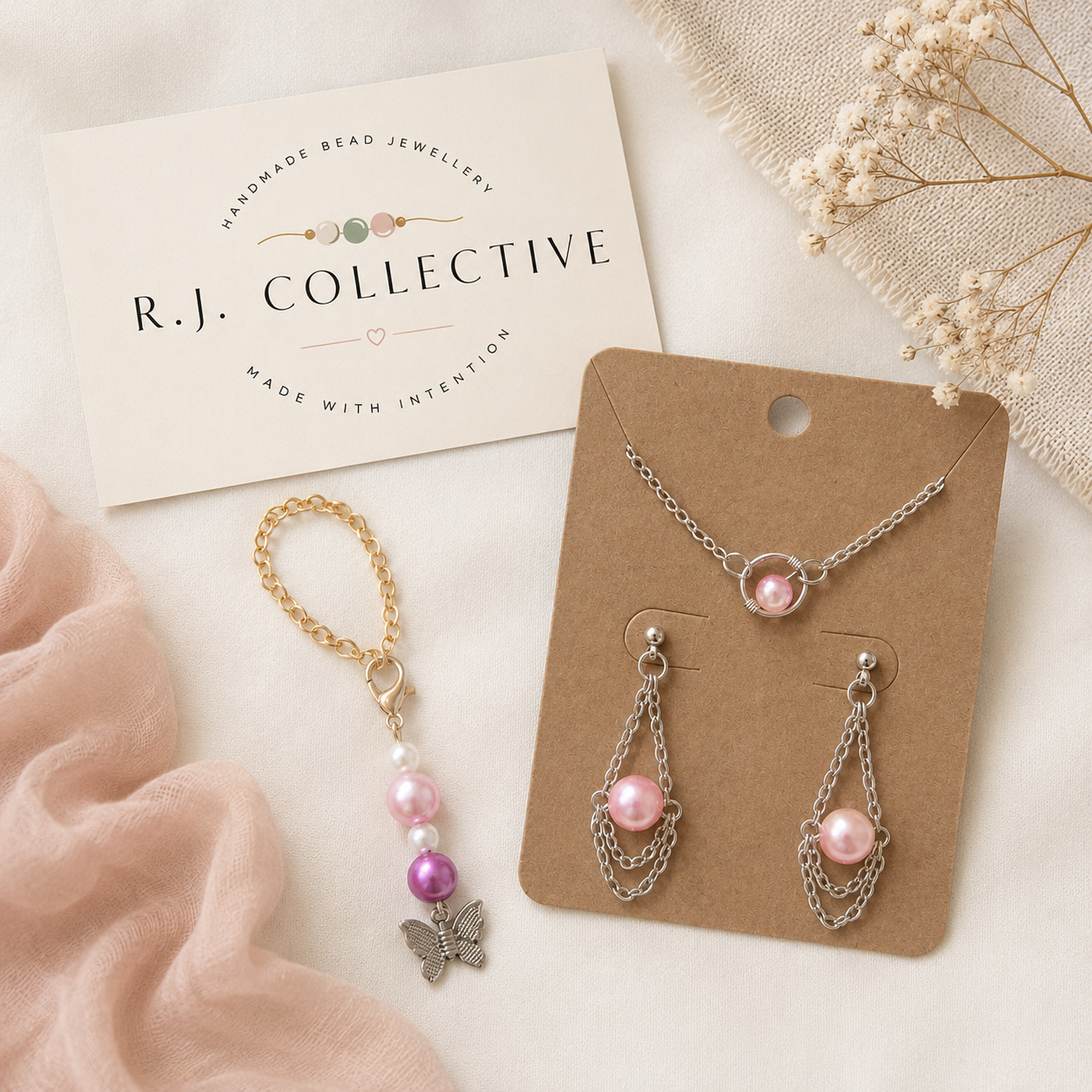 Jewelry set including a necklace with pink beads, earrings with chain and pink beads, and a bracelet with beads and a butterfly charm, displayed on brown cards, with a white card reading "R.J. Collective" and floral decorations.