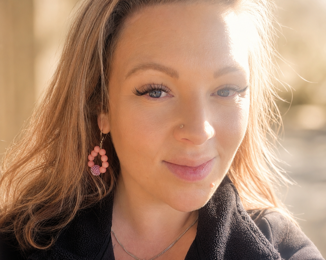 Close-up of a woman with light brown hair, wearing pink earrings, a nose piercing, and a black jacket, outdoors with sunlight on her face.