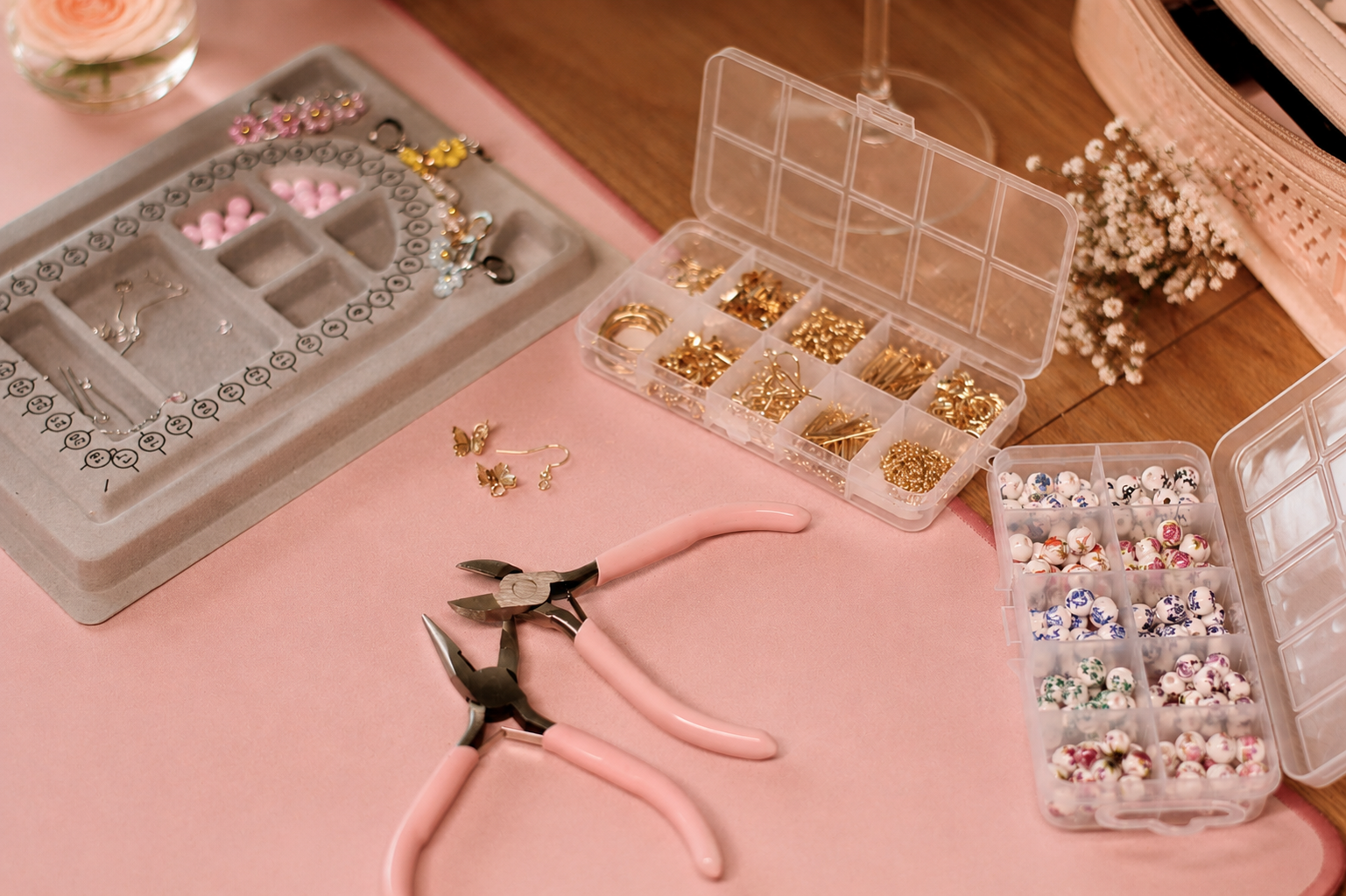 Jewelry-making supplies including jewelry pliers, earrings, rings, gold-colored charms, and assorted beads in various colors and designs, organized in plastic containers on a pink and wooden surface.