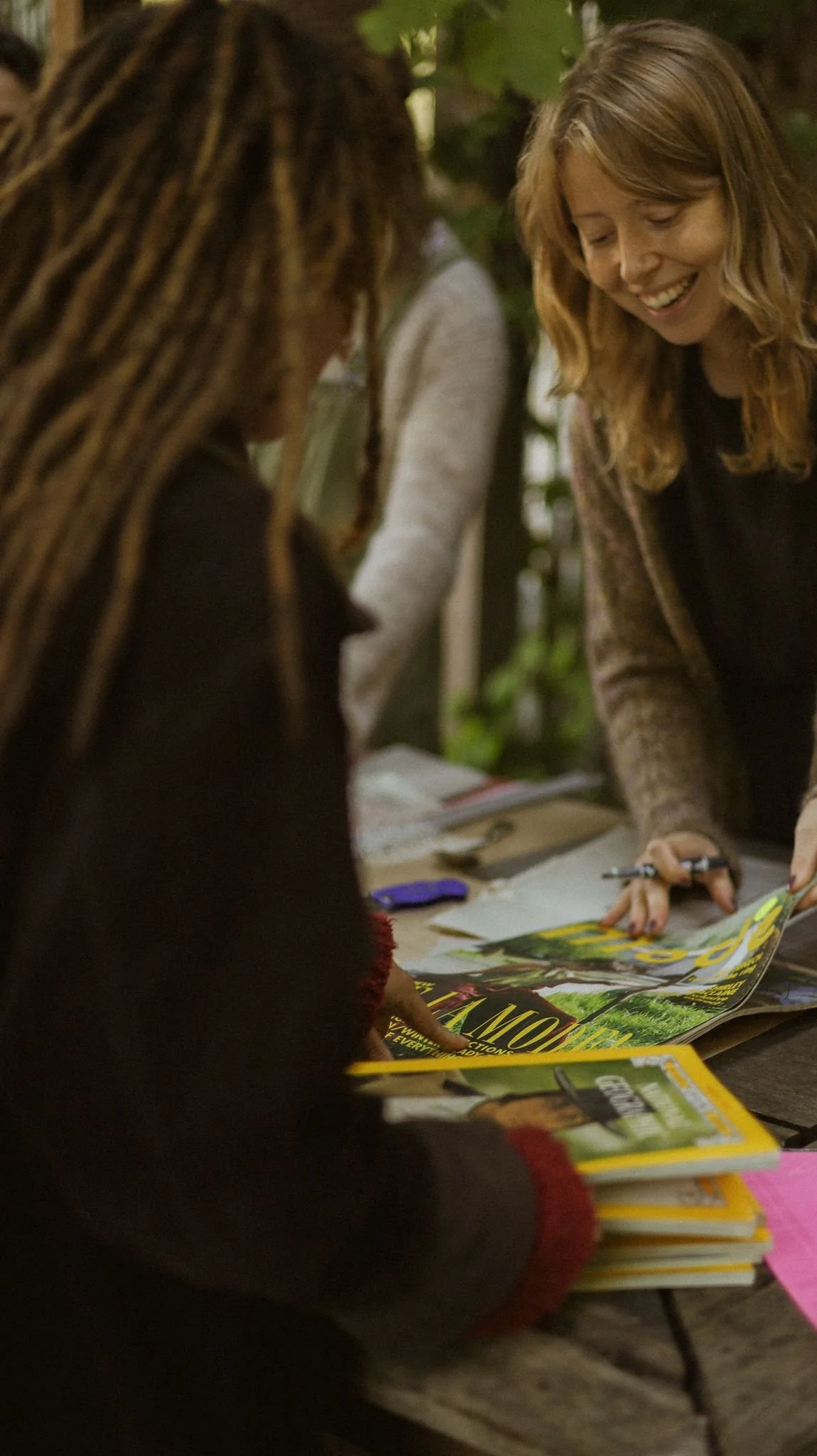 Two women at looking at zines on a table, with one woman smiling and holding a pen, and other magazines or books in the foreground.