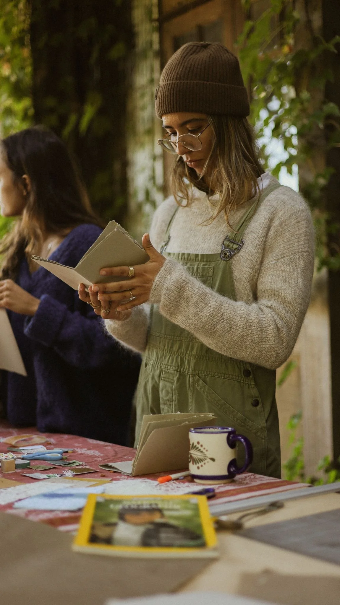 A woman wearing glasses, a brown beanie, and a green apron stands at a table, reading a notebook. There are craft supplies and photos on the table, and another person is partially visible in the background.