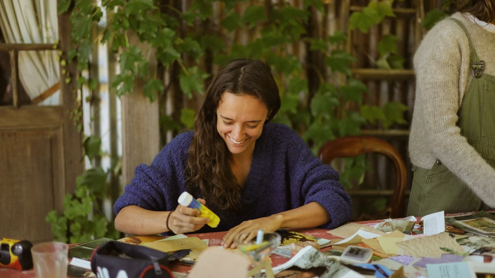 A woman with long curly brown hair smiling while sitting at a table with crafting supplies, holding a yellow glue stick in her right hand, in a room with green leafy plants in the background.