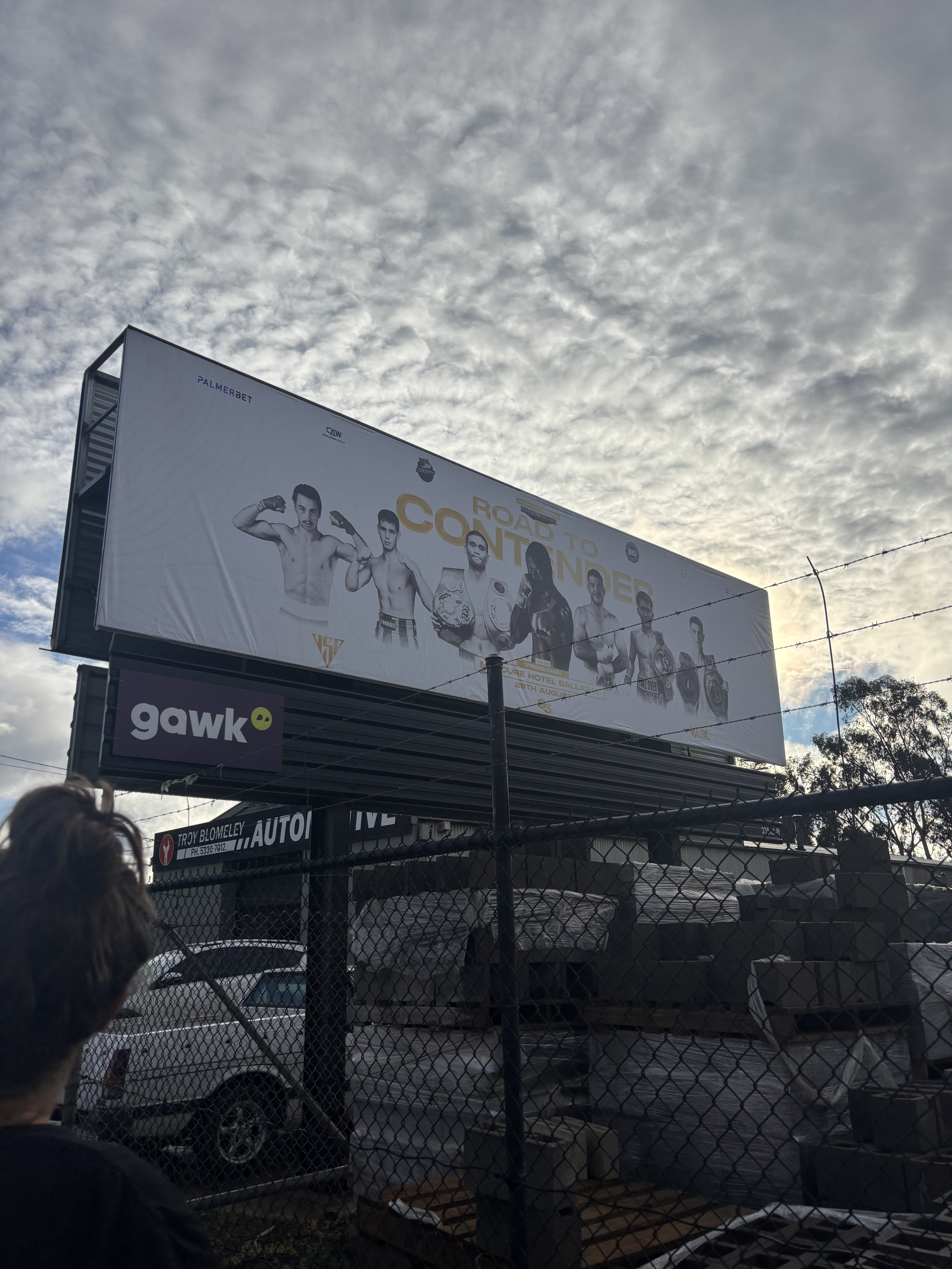 A large billboard advertisement featuring several boxing fans or fighters, with the words "Road to Contender" and logos for Palmerbet and other sponsors, against a cloudy sky background.