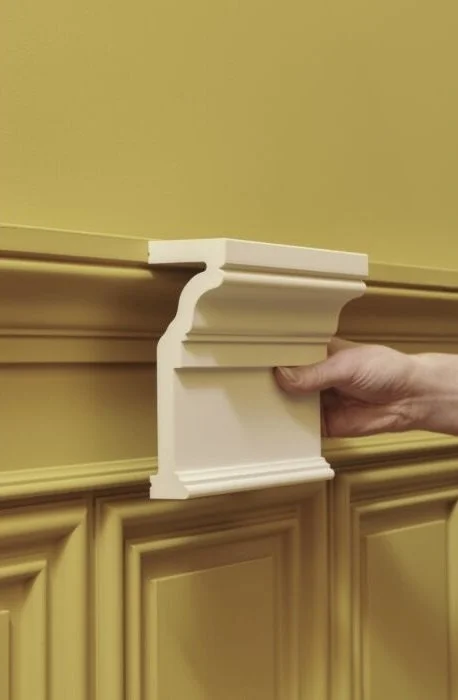 Mann hält eine weiße Zierleiste, die in eine Wandverkleidung integriert ist, vor einer gelben Wand mit geschnitztem Holzpanel.