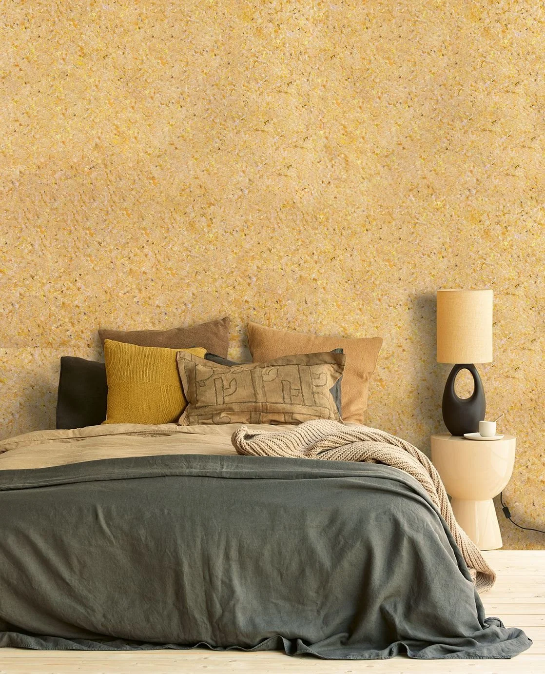 Schlafzimmer mit gemustertem gelblichem Wand und Bett mit dunklem und beigem Bettzeug, mehreren Kissen in Erdtönen, und einer beige Decke. Neben dem Bett ein runder Nachttisch mit schwarzer Oase-Lampe und einer weißen Kaffeetasse.