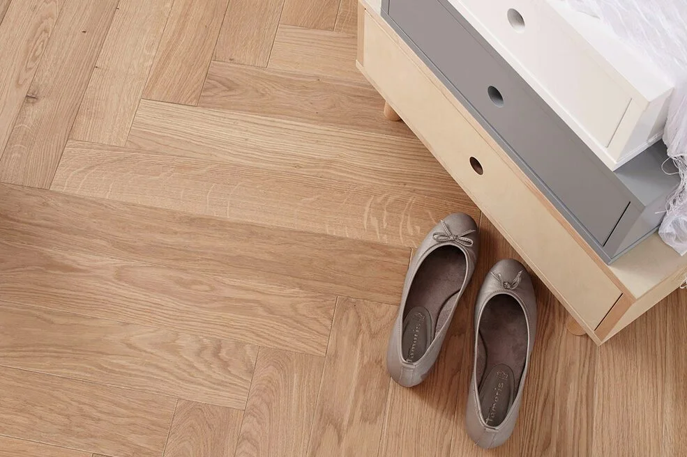 Ein Paar beige Ballerinas liegt auf einem Holzboden neben einer Kommode.