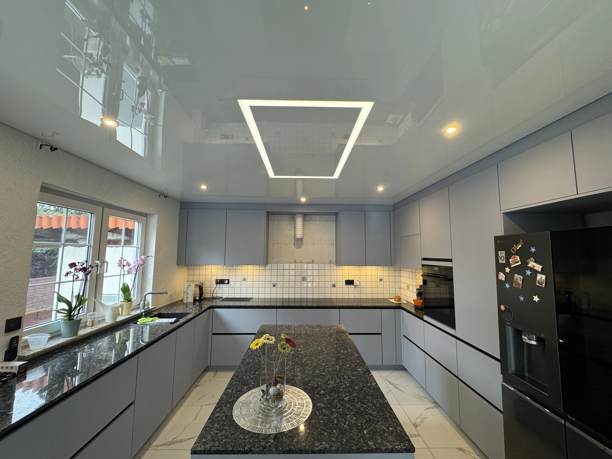 Moderne Küche mit schwarzen Granit-Arbeitsflächen, hellgrauen Schränken, großen Fenstern mit Pflanzen im Fensterbrett und einer Zentraltisch mit Blumen, weiße Wandfliesen und eine Deckenleuchte in Form eines rechteckigen Rahmens.