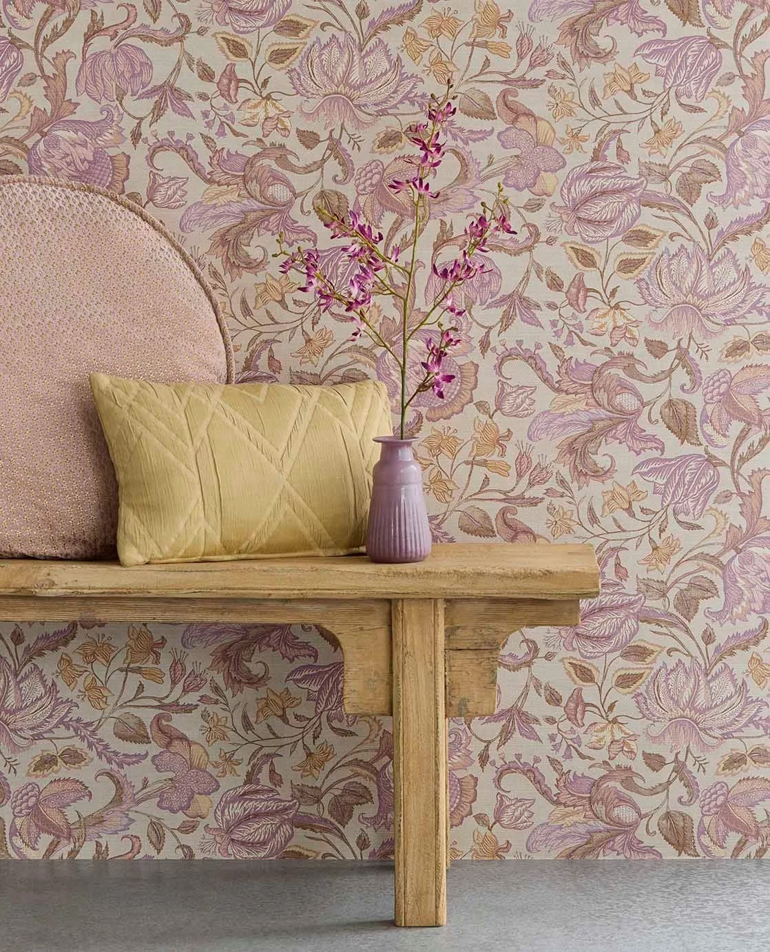 Vase mit pinken Blumen auf Holzbank vor einer Wand mit floral gemusterter Tapete in Pastellfarben.