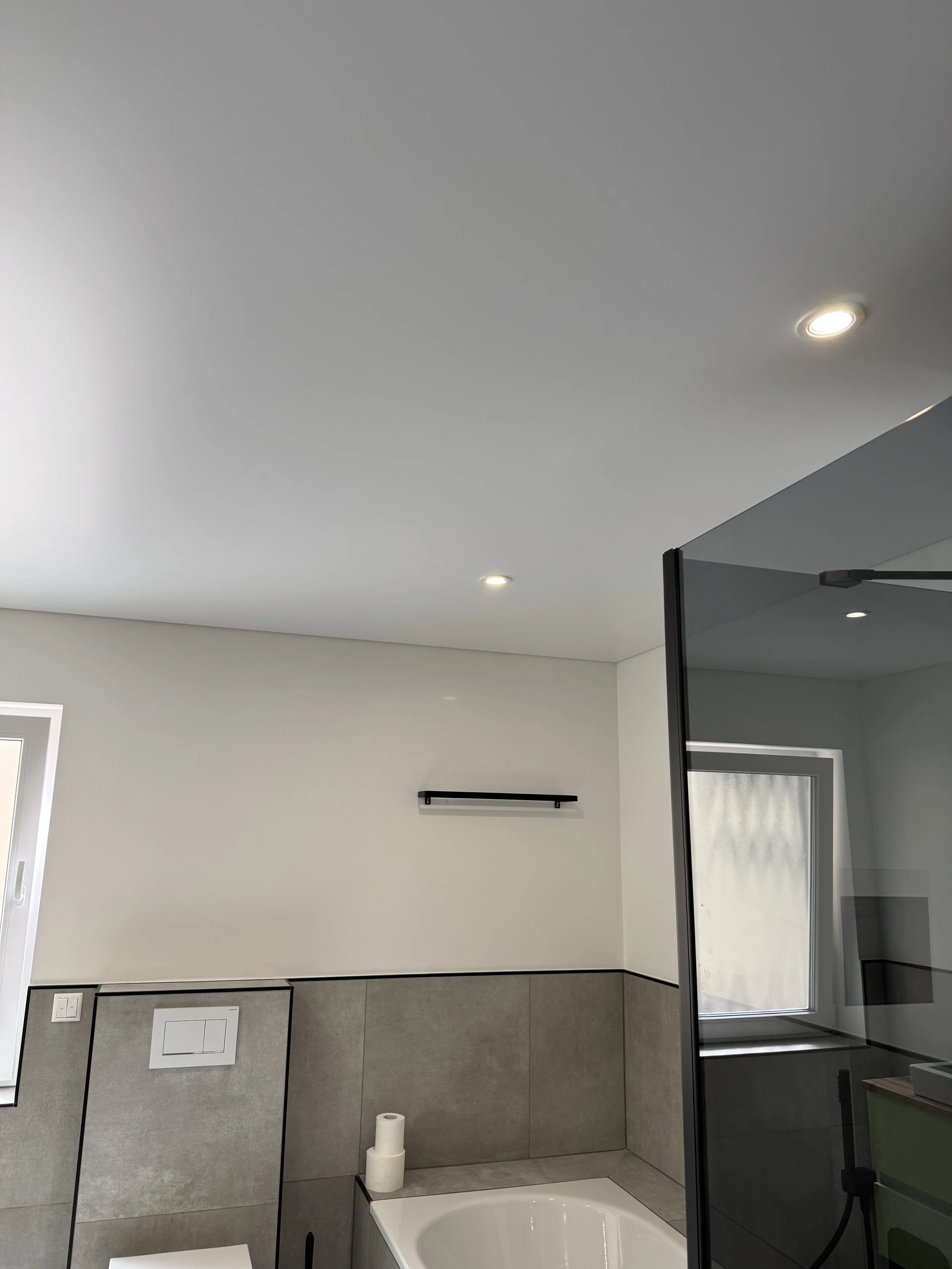 Ein modernes Badezimmer mit einer Badewanne, grauen Wandfliesen, einer minimalistisch gestalteten Wand mit verstecktem Waschbecken, einem kleinen Fenster und einer schwarzen Zierleiste.
