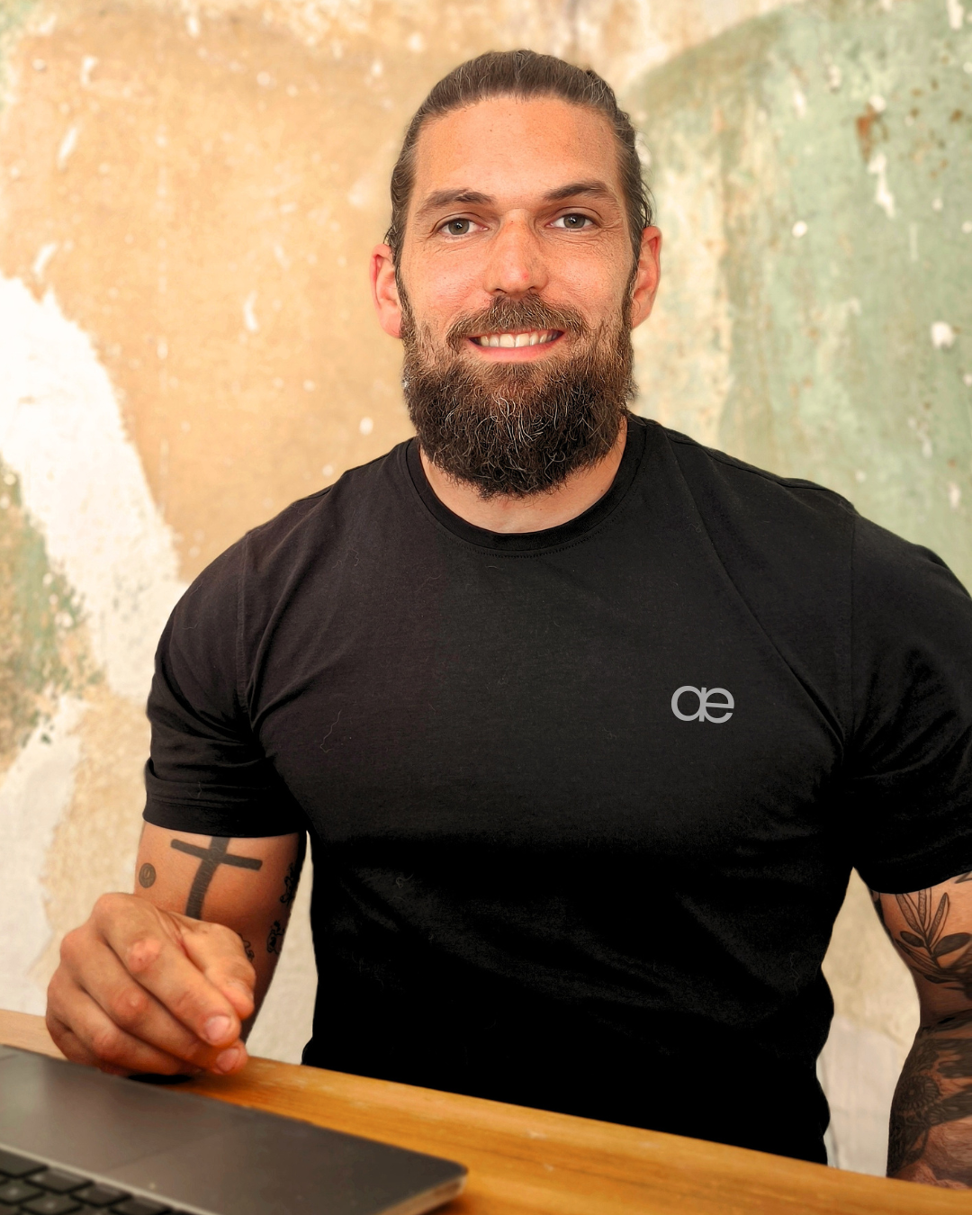 Ein Mann mit dunklem Haar und Bart, trägt ein schwarzes T-Shirt mit einem kleinen Logo, sitzt an einem Tisch mit einem Laptop, im Hintergrund eine bunte Wand.