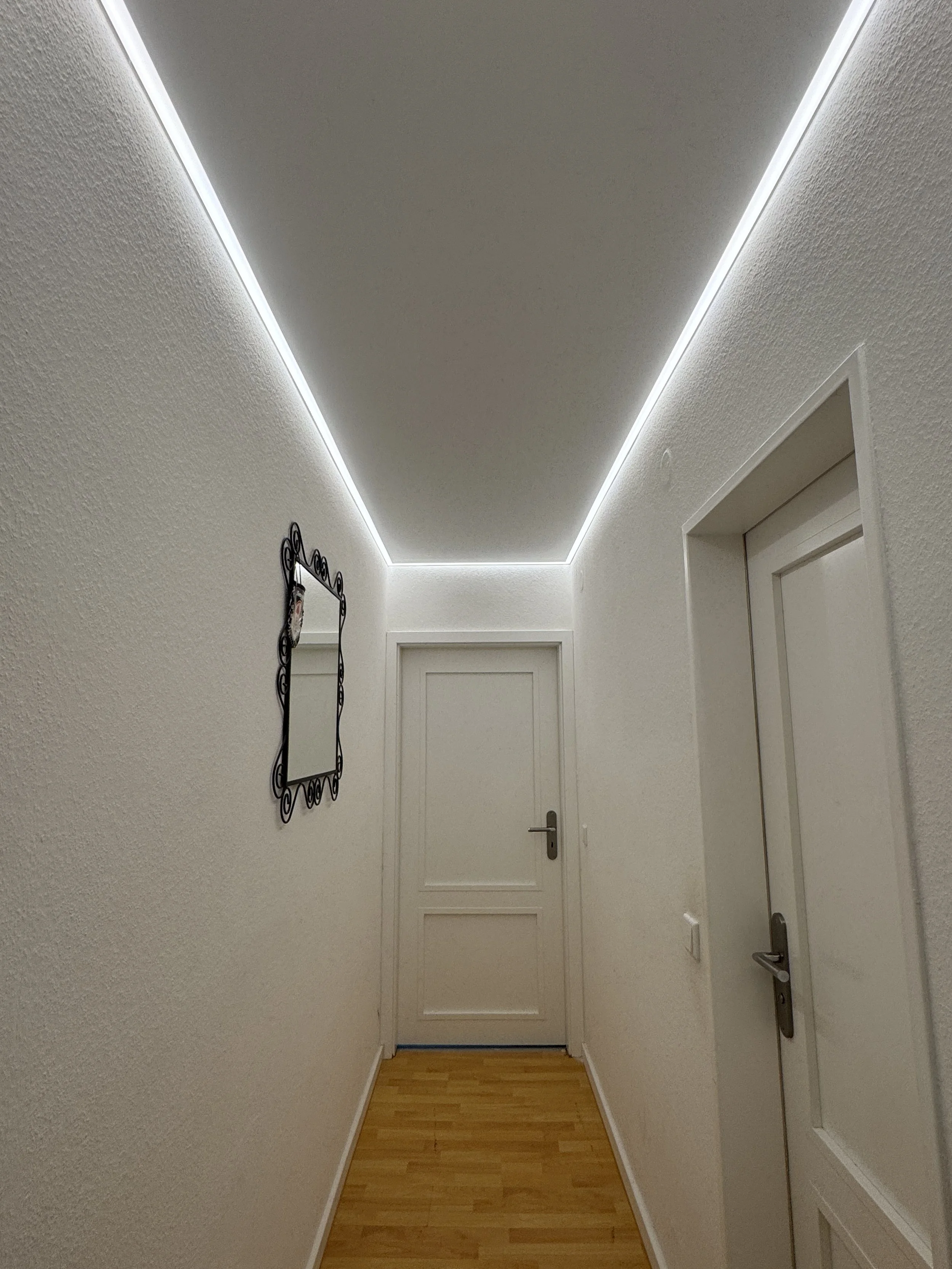 Ein schmaler Flur mit weißen Wänden, Holzboden und einer Tür an der Wand auf der rechten Seite. An der linken Wand hängt ein dekorativer Spiegel.