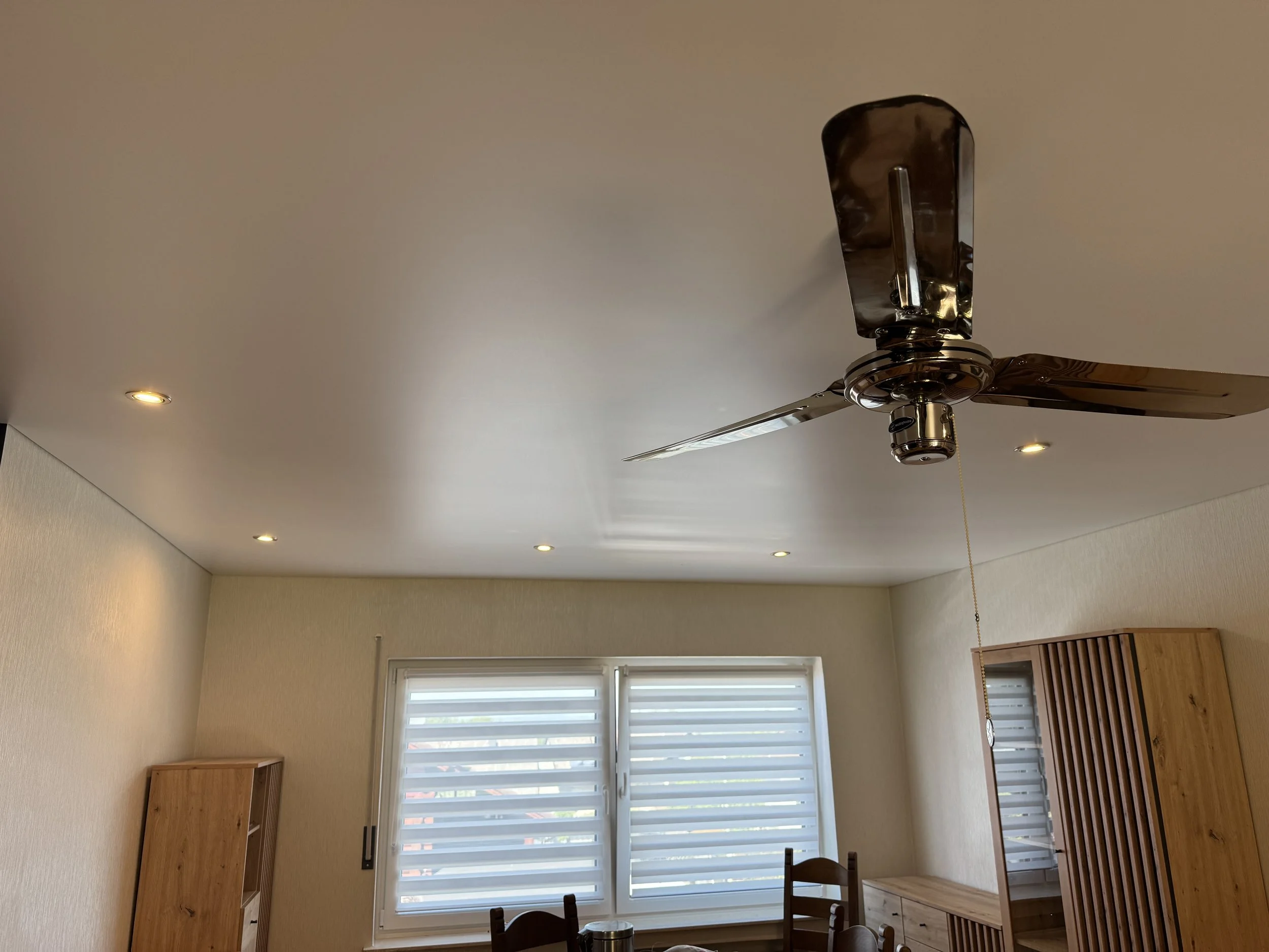Ein Deckenventilator mit drei Metallflügeln an einer weißen Zimmerdecke, umgeben von mehreren Deckenleuchten. Ein Fenster mit weißen Lamellen ist im Hintergrund sichtbar.