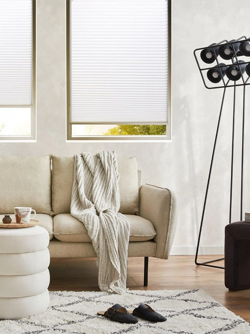 Helles Wohnzimmer mit Sofa, weißen Fensterjalousien, gestrickter Decke, Teppich, Paar schwarze Hausschuhe, Stehlampe und kleine Deko-Artikel auf einem Tisch.
