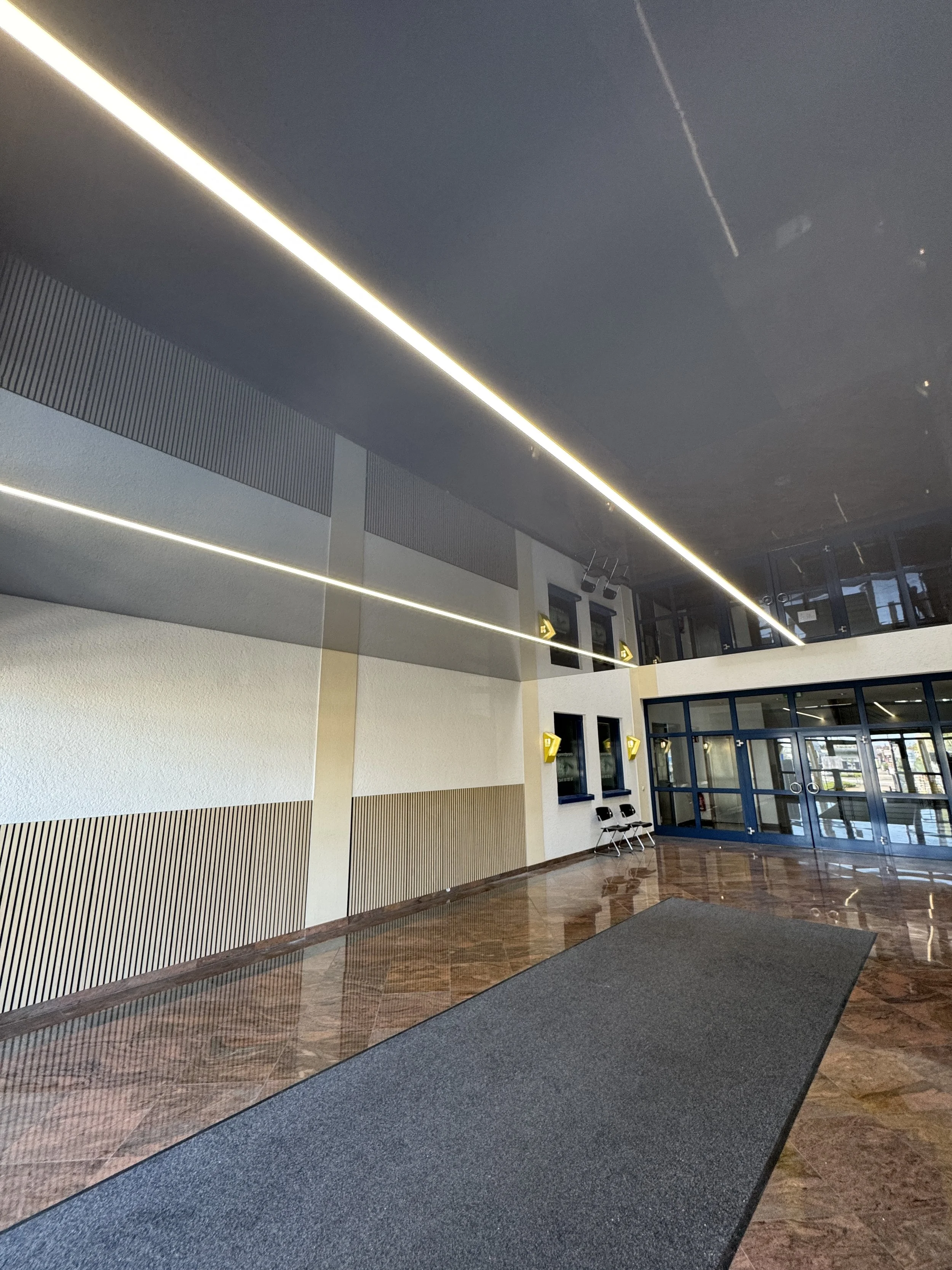 Inneneingang eines modernen Gebäudes mit großen Glasflügeltüren, Sitzbank, dunklem Bodenbelag und Deckenbeleuchtung.
