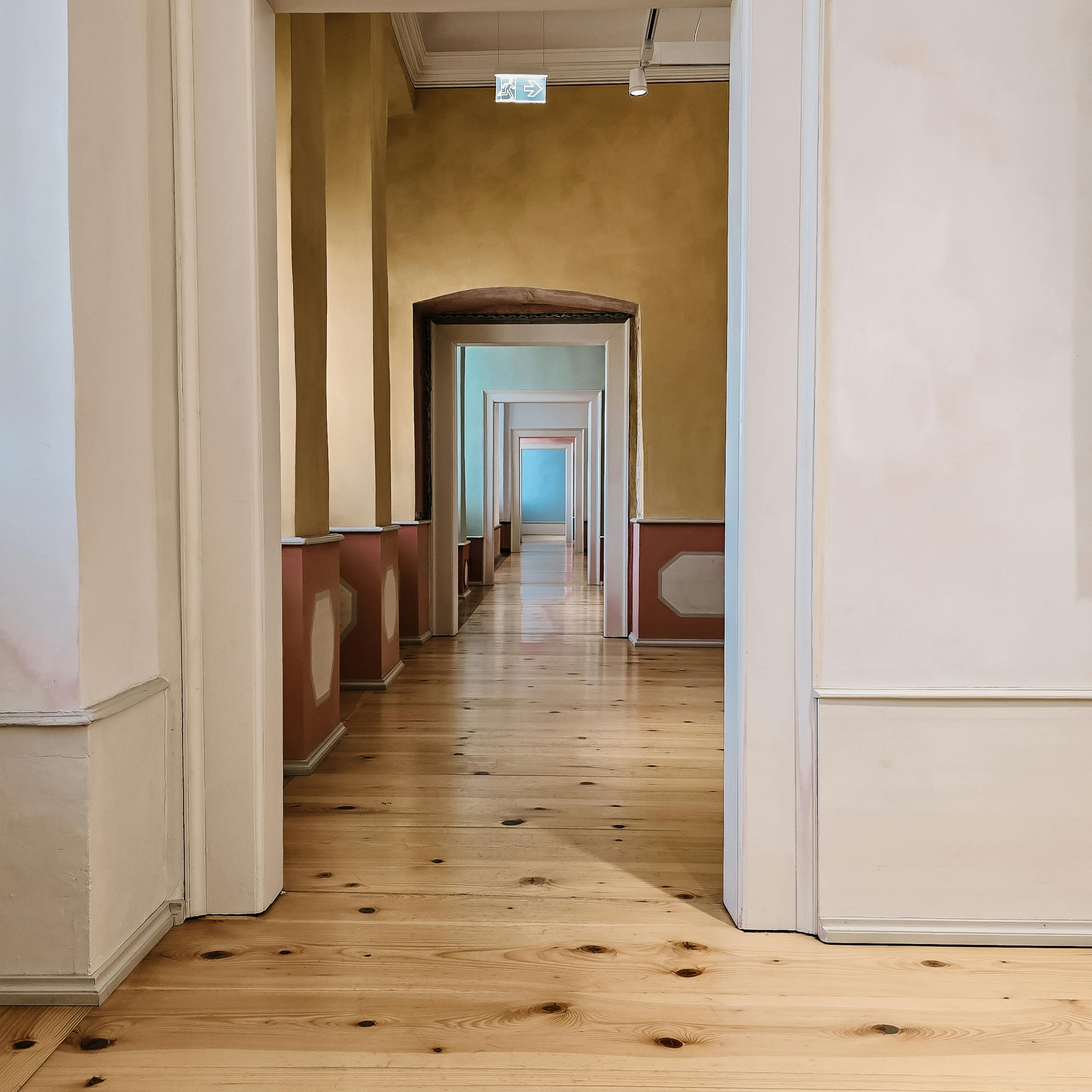Mehrere Türen in einem Korridor, Blick nach hinten, Holzfußboden, weiße Wände, Decke mit Lüftung und Leuchte, Notausgangsschild hängt von der Decke.