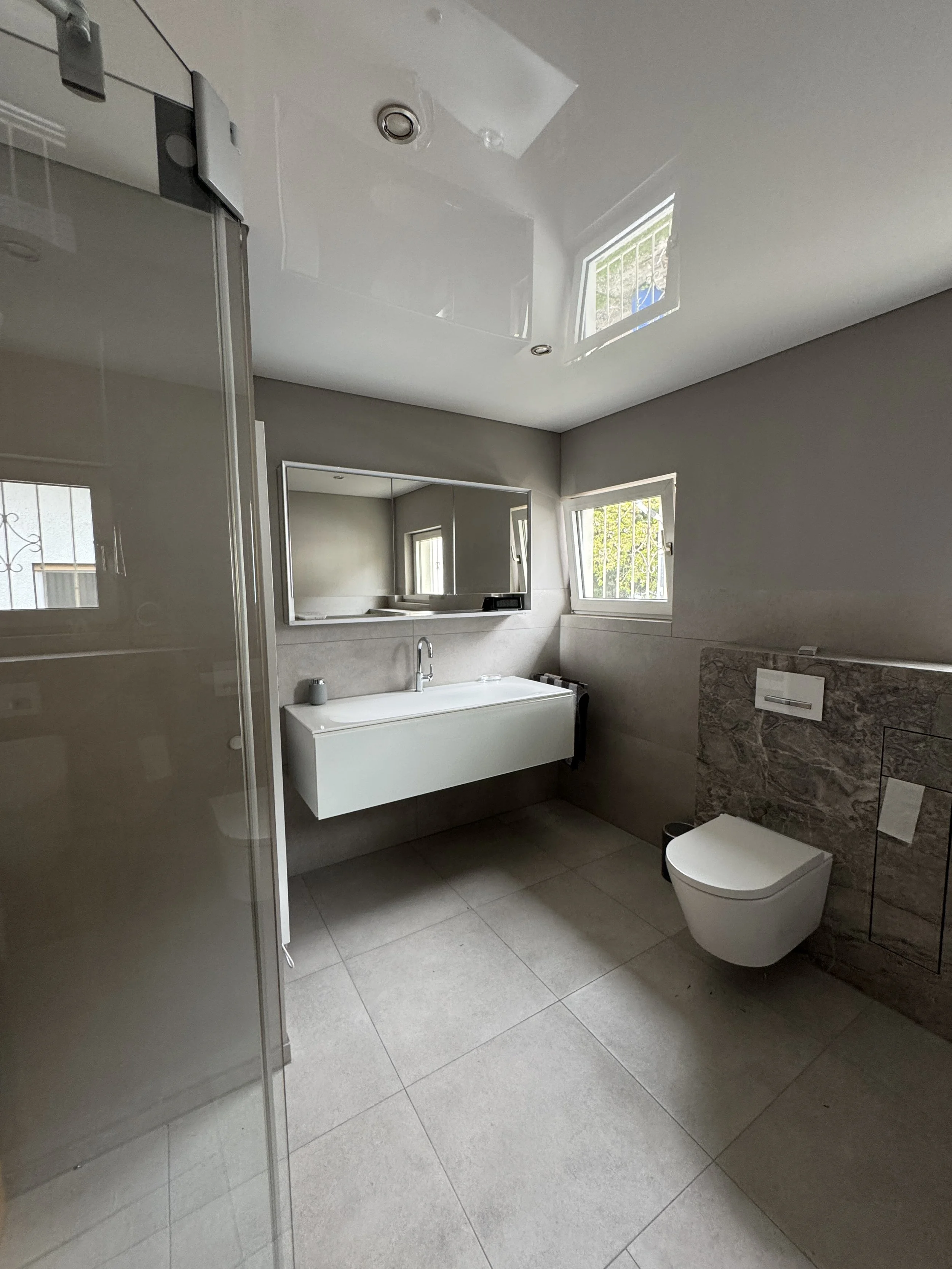 Moderner Badezimmer mit einer weißen Wandwaschbecken, Spiegel, Toilette, Fenster und Fliesenboden.